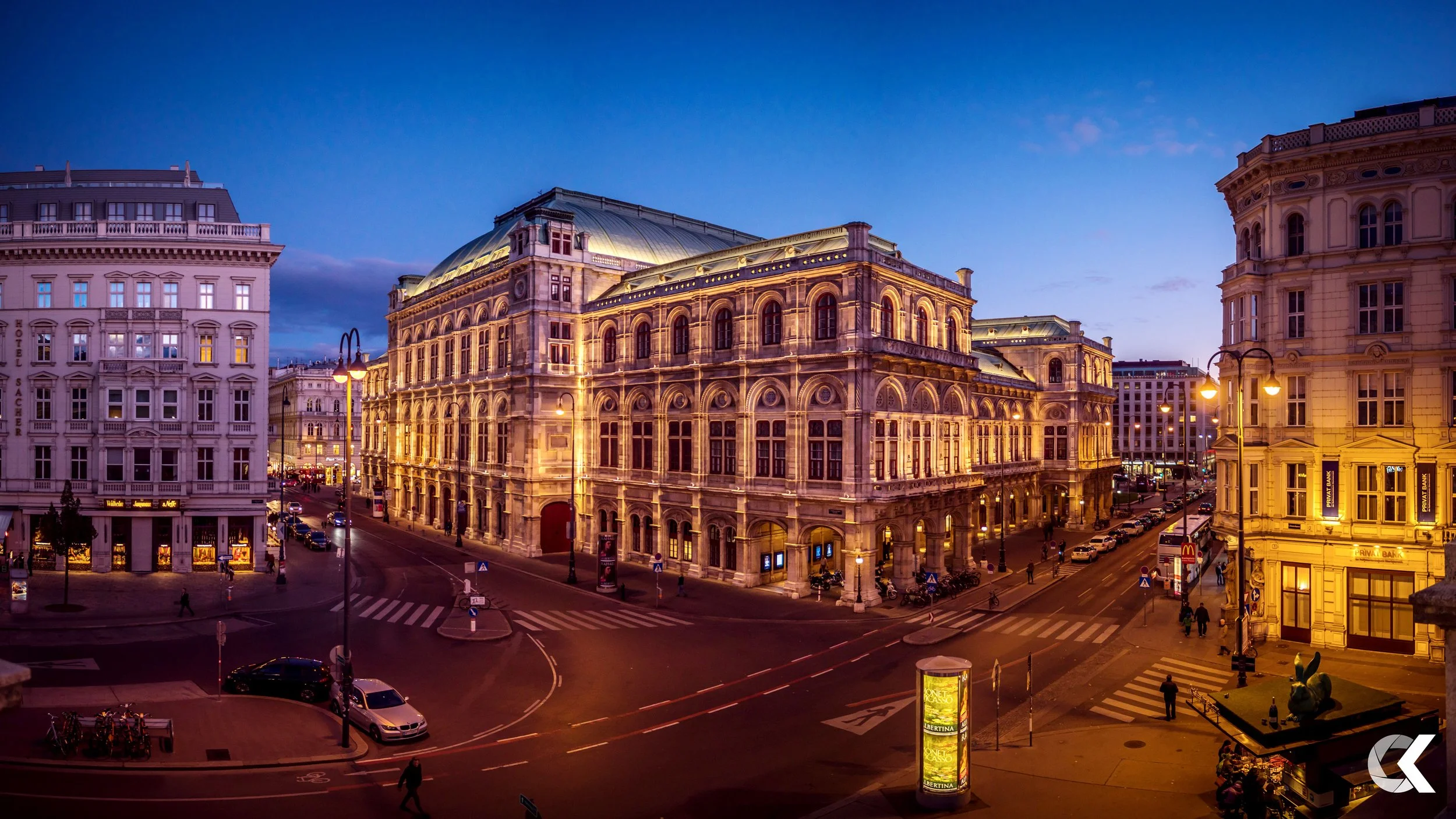 Nighttime city scene with historic buildings illuminated, people walking on sidewalks, cars on the streets, and a digital billboard in front.