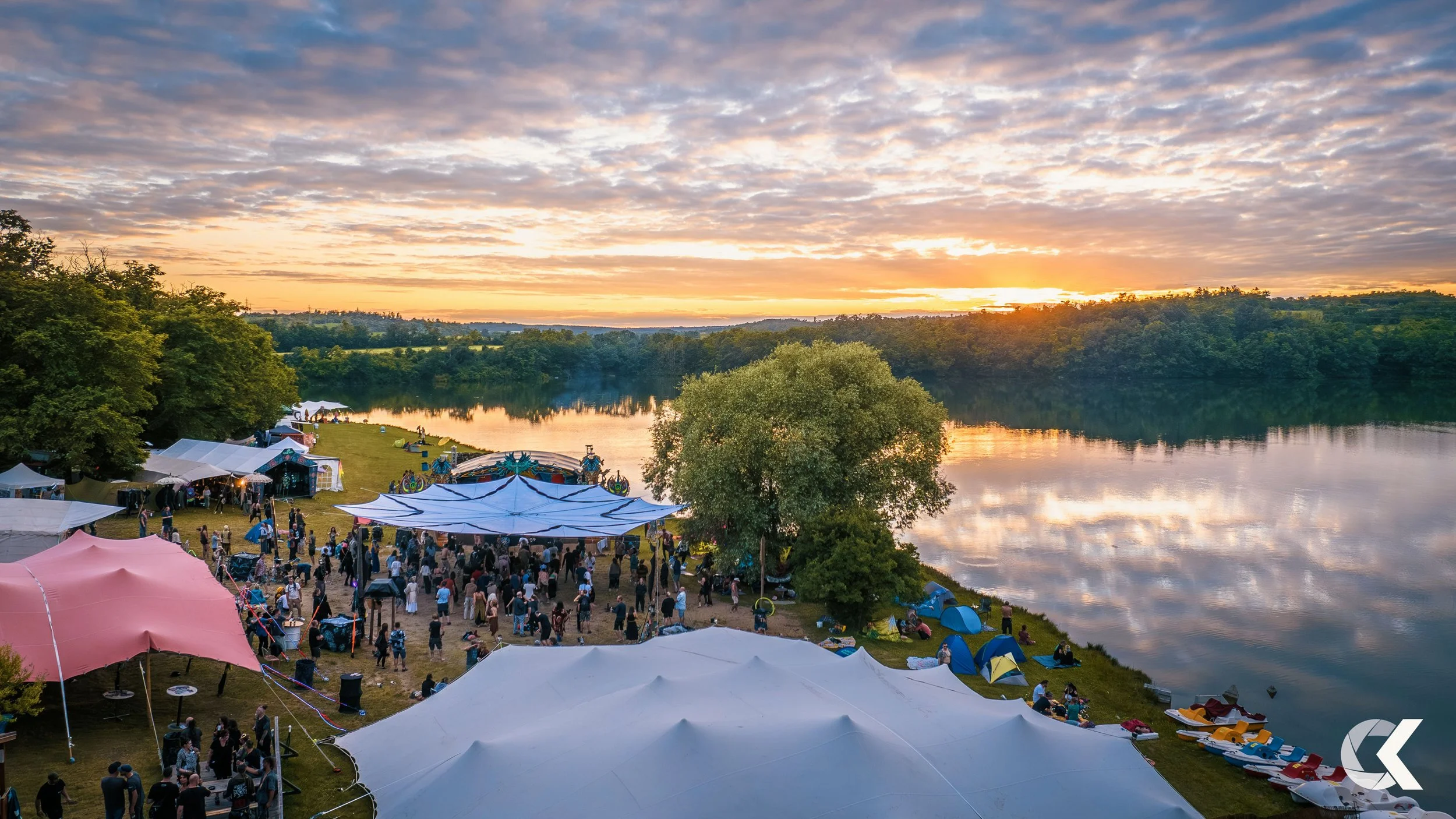 A lively outdoor event near a body of water at sunset, with tents, boats, and many people enjoying various activities.