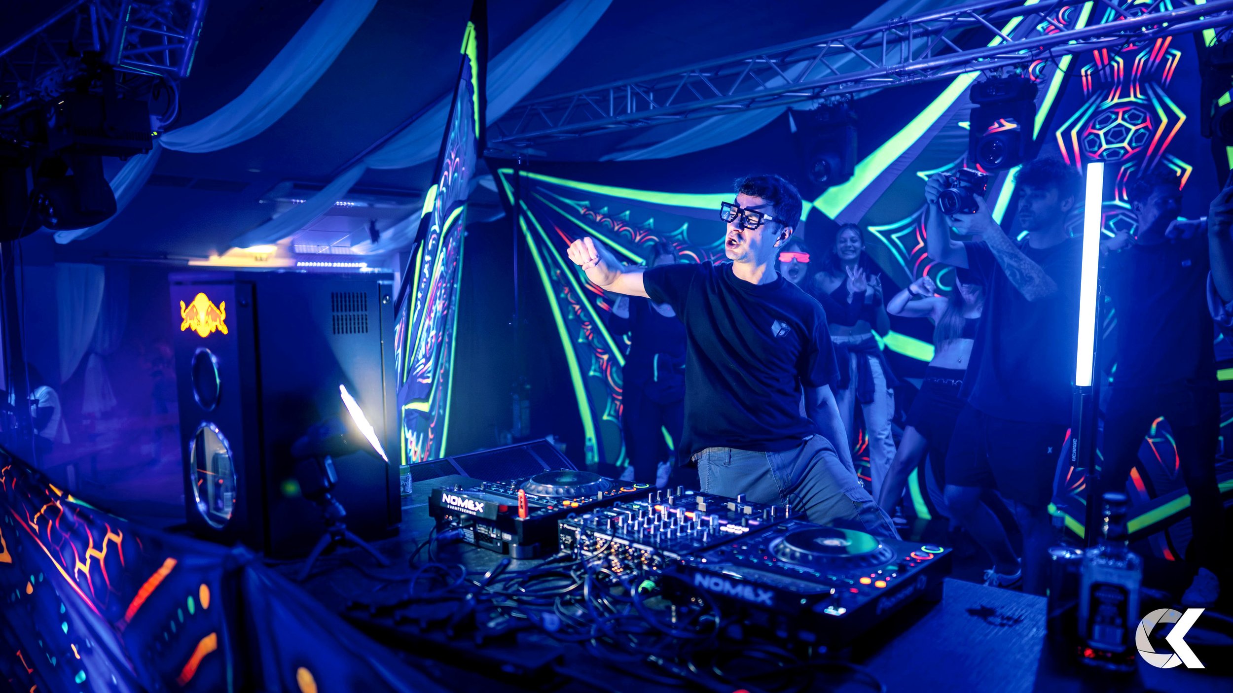 DJ performing at a neon-lit nightclub with people dancing and colorful geometric light patterns in the background.