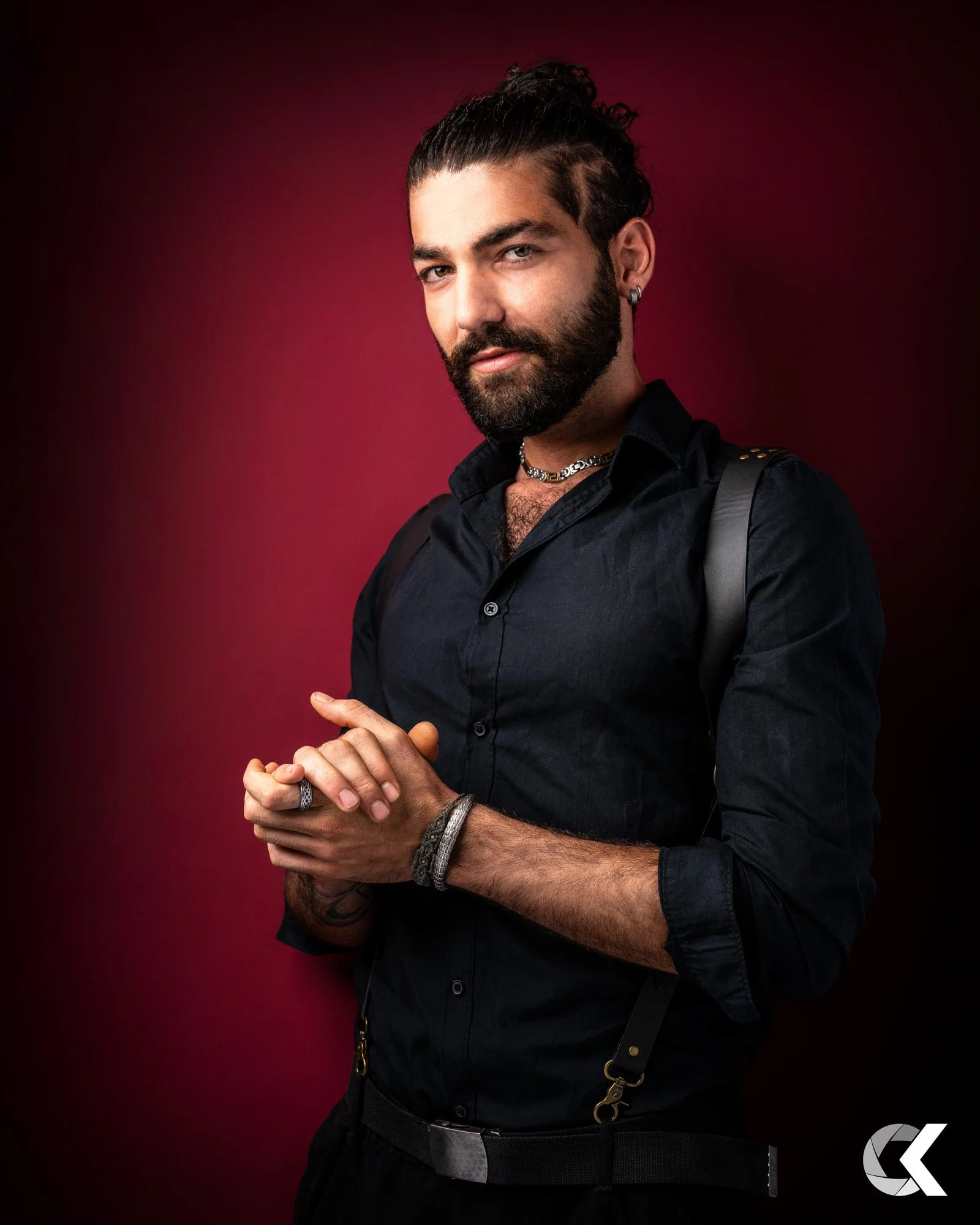 Ein junger Mann mit dunklem Haar, Bart und Piercing trägt schwarze Kleidung, eine Halskette und Armbänder, steht vor einem rot-schwarzem Hintergrund, schaut direkt in die Kamera, wirkt selbstbewusst.