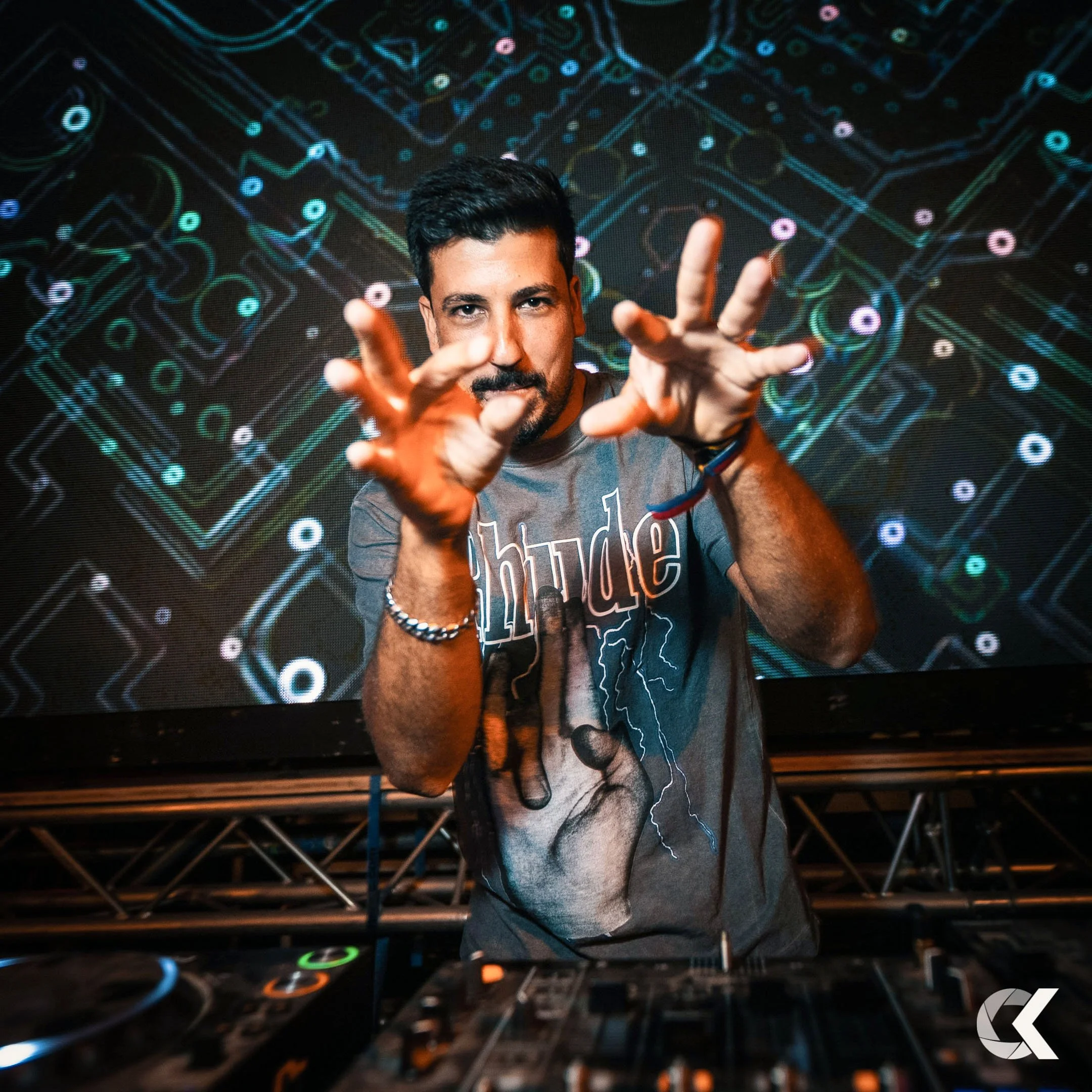 A DJ performing in front of a digital circuit board background, wearing a graphic t-shirt, with hands raised and reaching toward the camera.