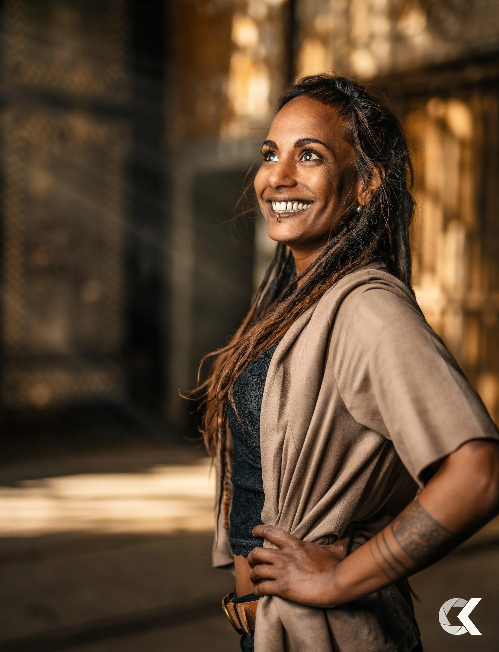 A woman with long dreadlocks and a nose piercing smiling and looking to the side, standing indoors with blurred warm background.