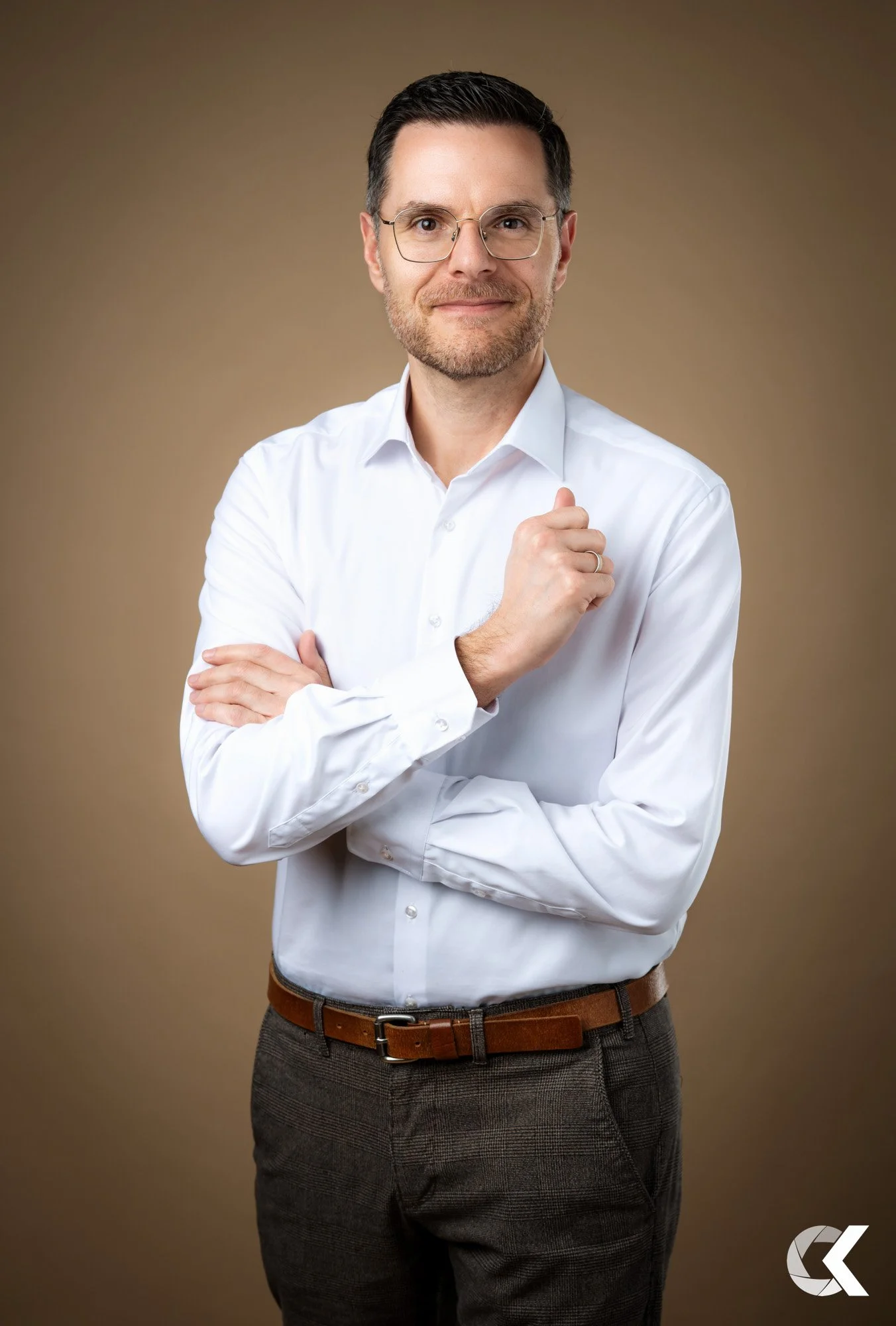A man with glasses and a beard in a white dress shirt standing against a brown background, with his arms crossed and looking at the camera.