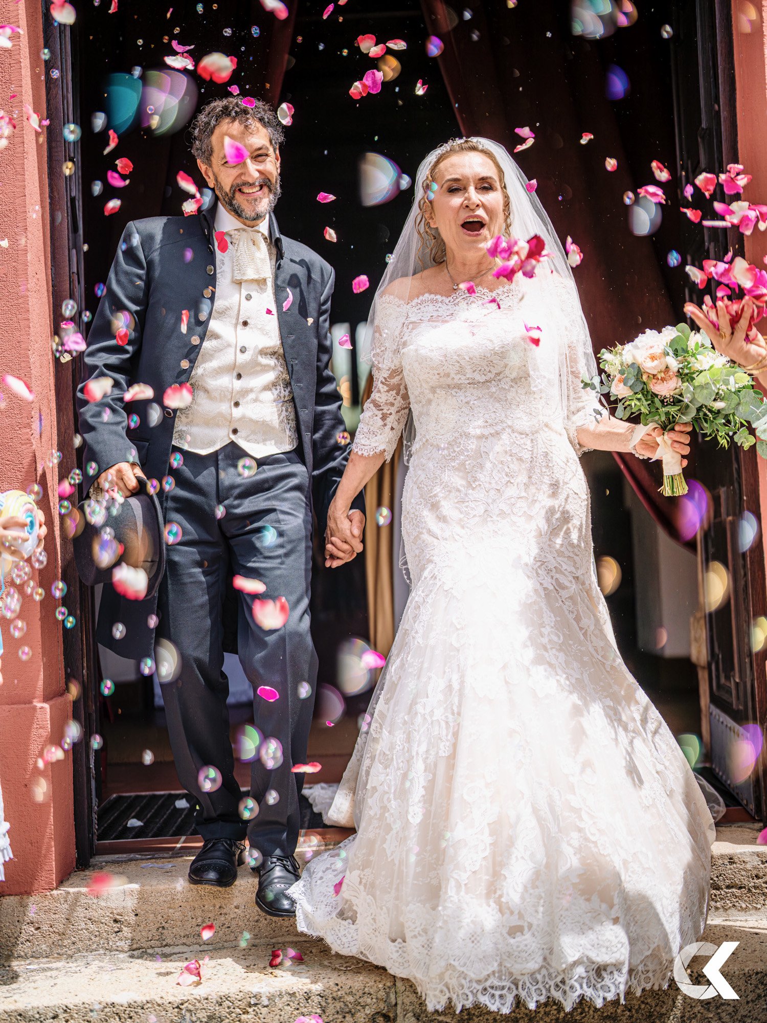 A bride and groom exiting a building, holding hands, with rose petals being thrown in celebration.
