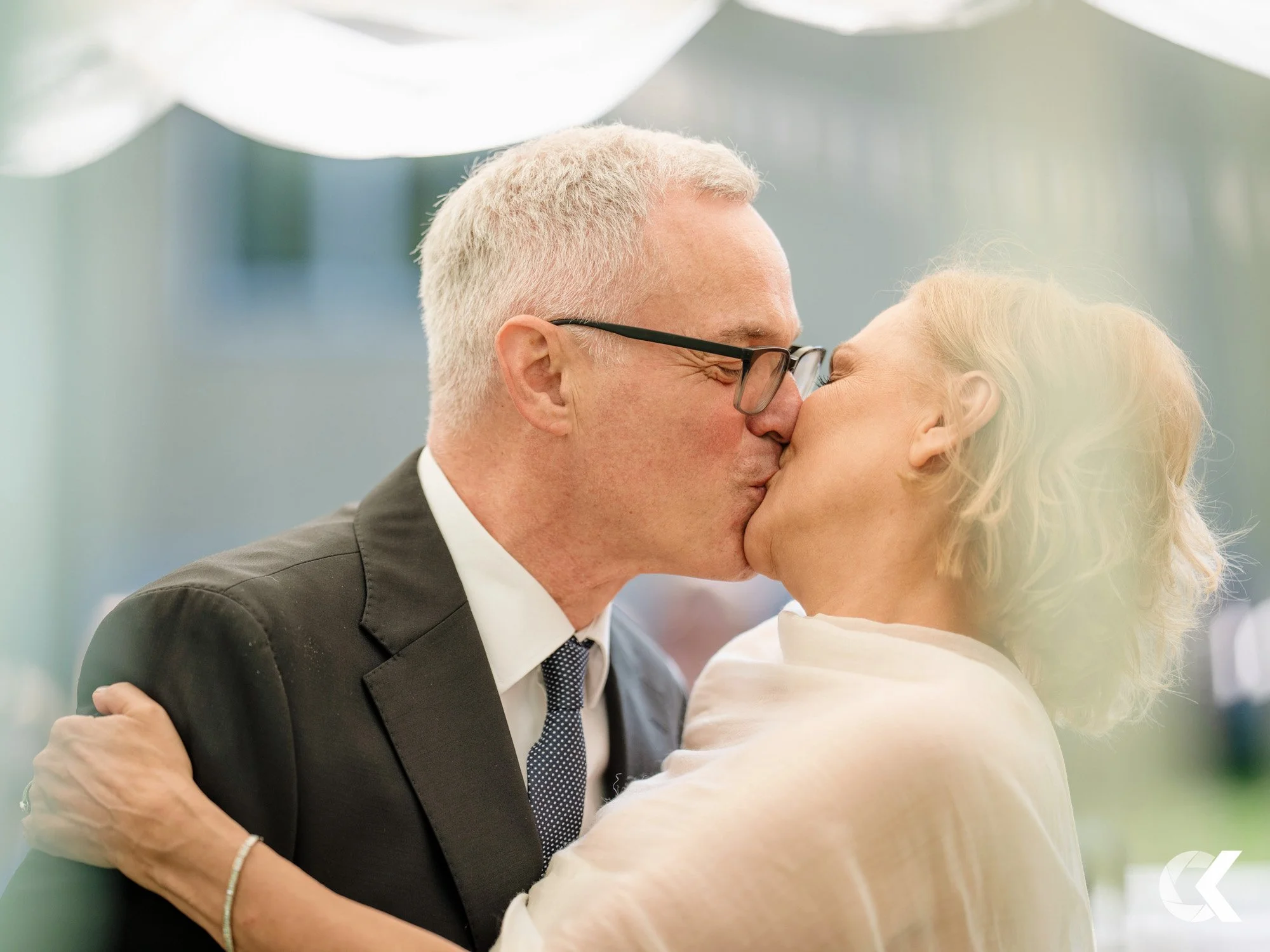 A couple is sharing a kiss, with the man wearing glasses and a suit, and the woman having short blonde hair and a cream-colored shawl.