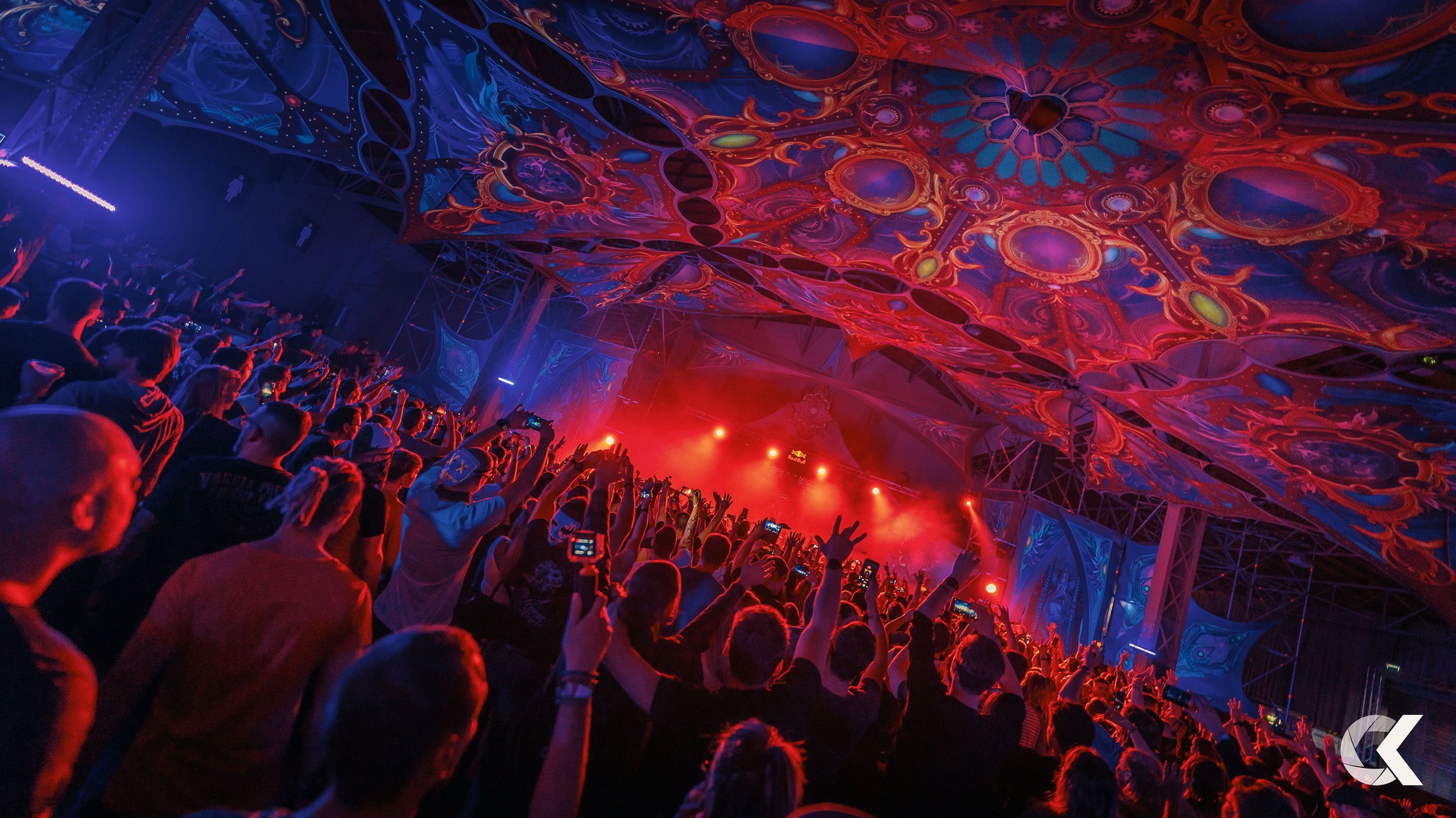 A crowded concert or music festival with people dancing and taking pictures under a colorful, psychedelic ceiling with vibrant patterns and stage lighting.