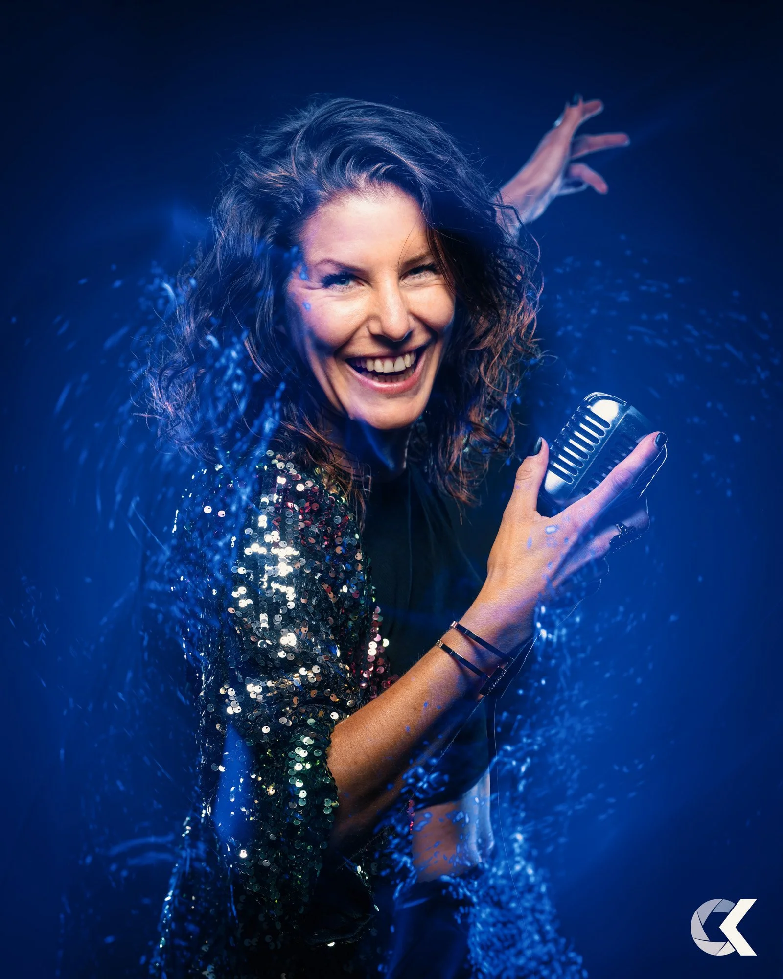 Eine lachende Frau mit dunklen, gelockten Haaren singt in einem Mikrofon, umgeben von blauen Lichteffekten und Glitzer, die eine energische Performance anzeigen.