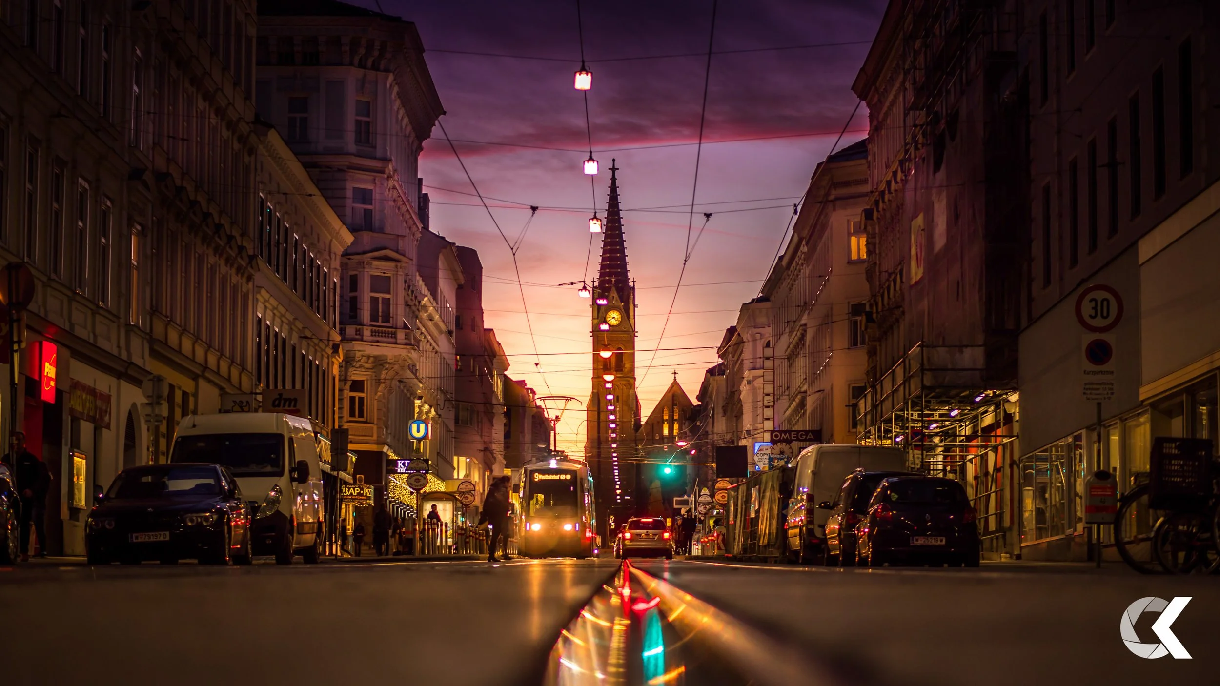 City street at dusk with tram approaching, streetlights and shop signs illuminated, in front of a church with a tall spire, reflecting on wet pavement.