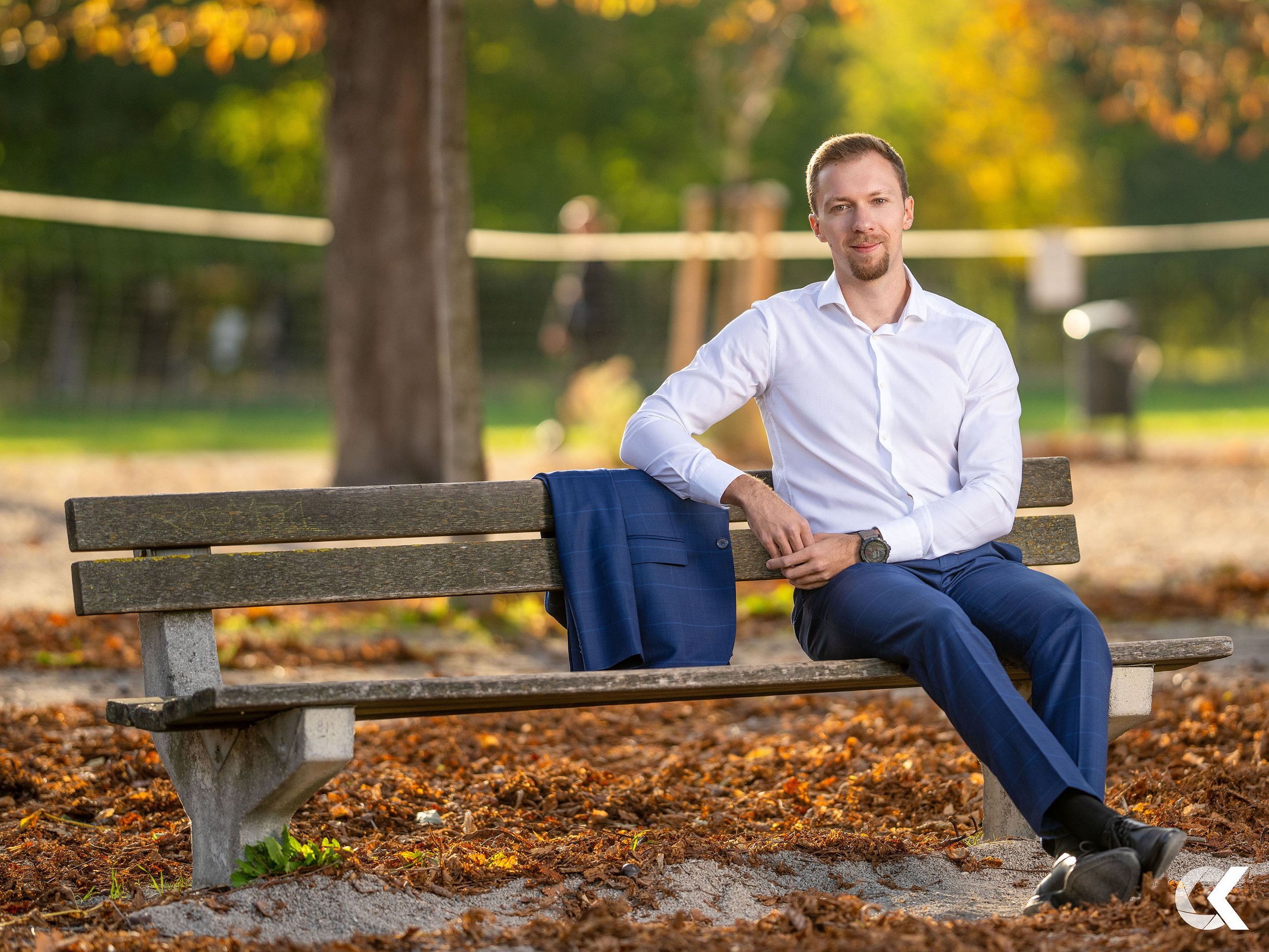 A man in a white dress shirt and blue suit pants sitting on a park bench outdoors in autumn. A blue suit jacket is draped over the back of the bench. The man is looking at the camera with a slight smile, and there are trees with green and orange leav