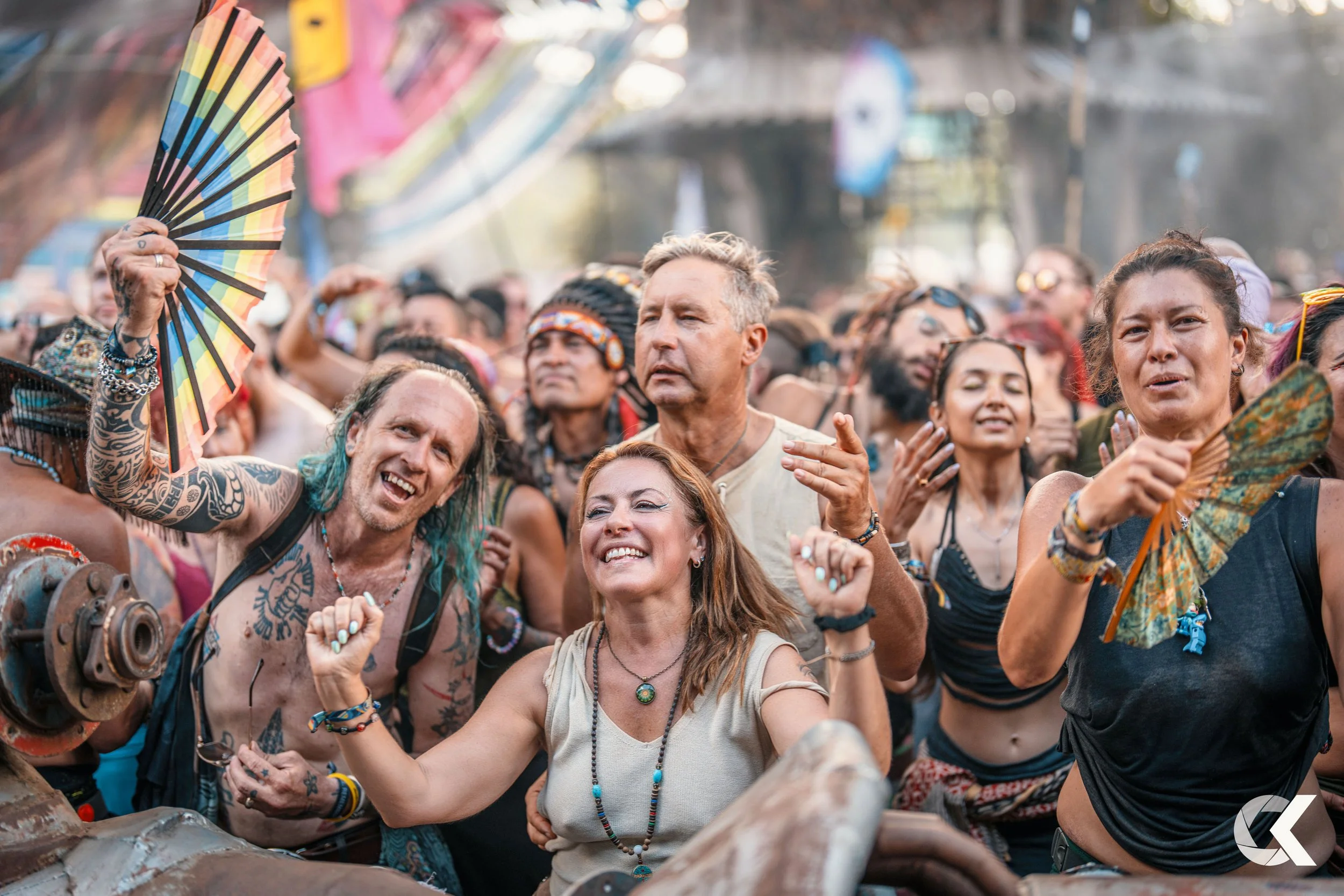 A group of diverse people enjoying a lively outdoor event or festival, dancing enthusiastically, with some holding colorful fans, surrounded by a crowd under sunny weather.