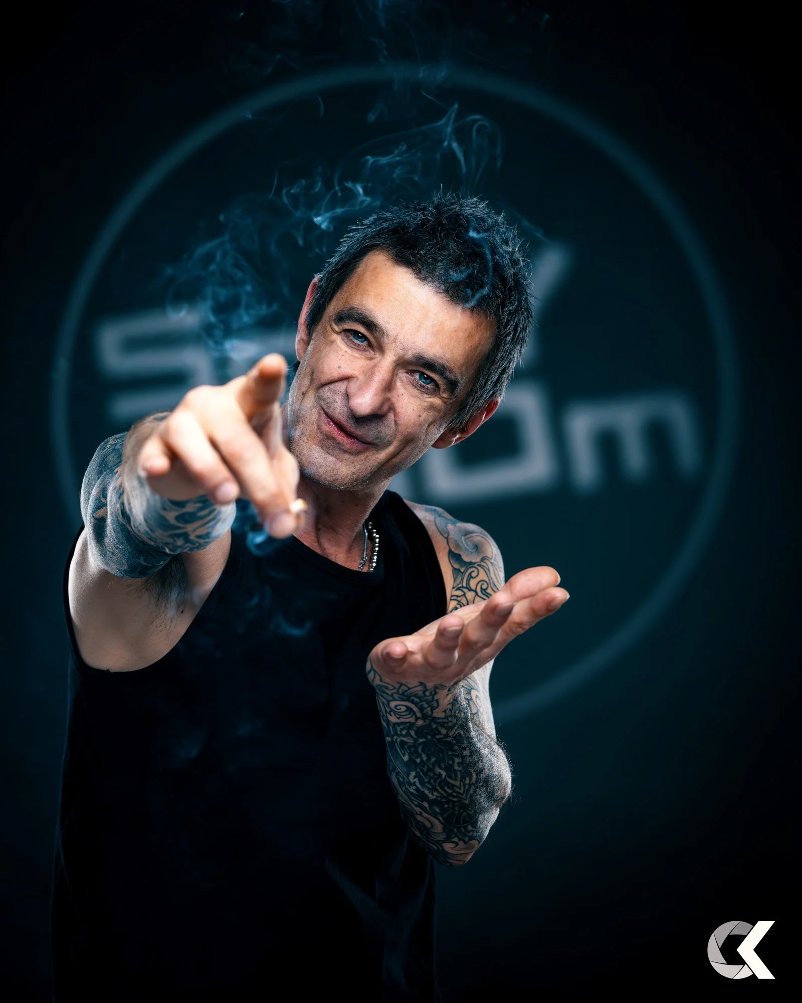 Man with black hair and tattoos on arms, wearing a black sleeveless shirt, pointing towards the camera with a confident expression, smoke around him, dark background with a blurred logo.