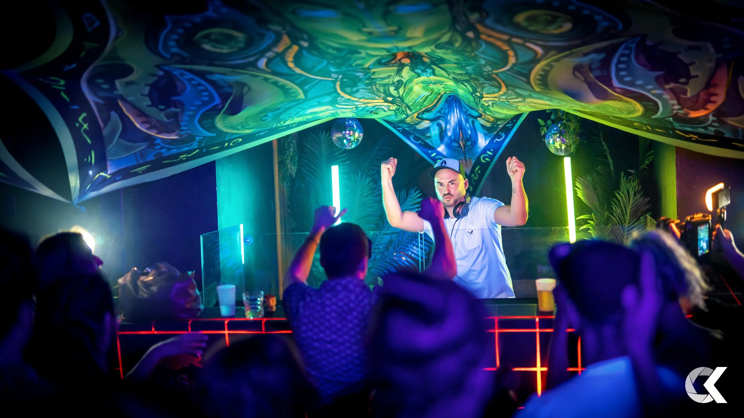 A DJ performing at a nightclub with colorful lighting and a psychedelic ceiling. The DJ is wearing a white t-shirt and a cap, raising both arms, while the crowd dances and takes photos.
