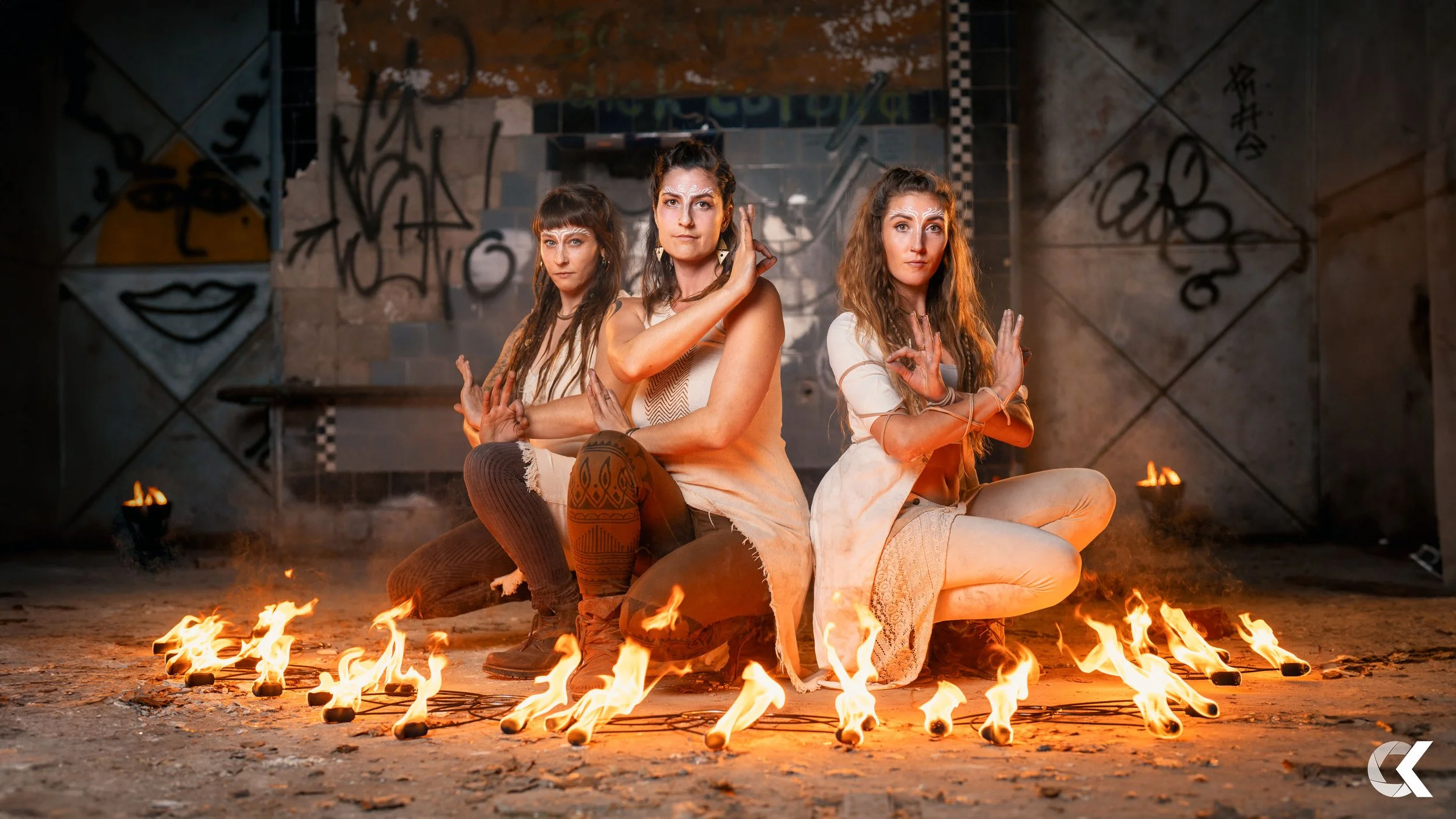 Three women dressed in bohemian style clothing posing in an abandoned industrial space with graffiti on the walls. They are sitting cross-legged on the ground surrounded by lit torches. The women have their hands positioned in various gestures, and t