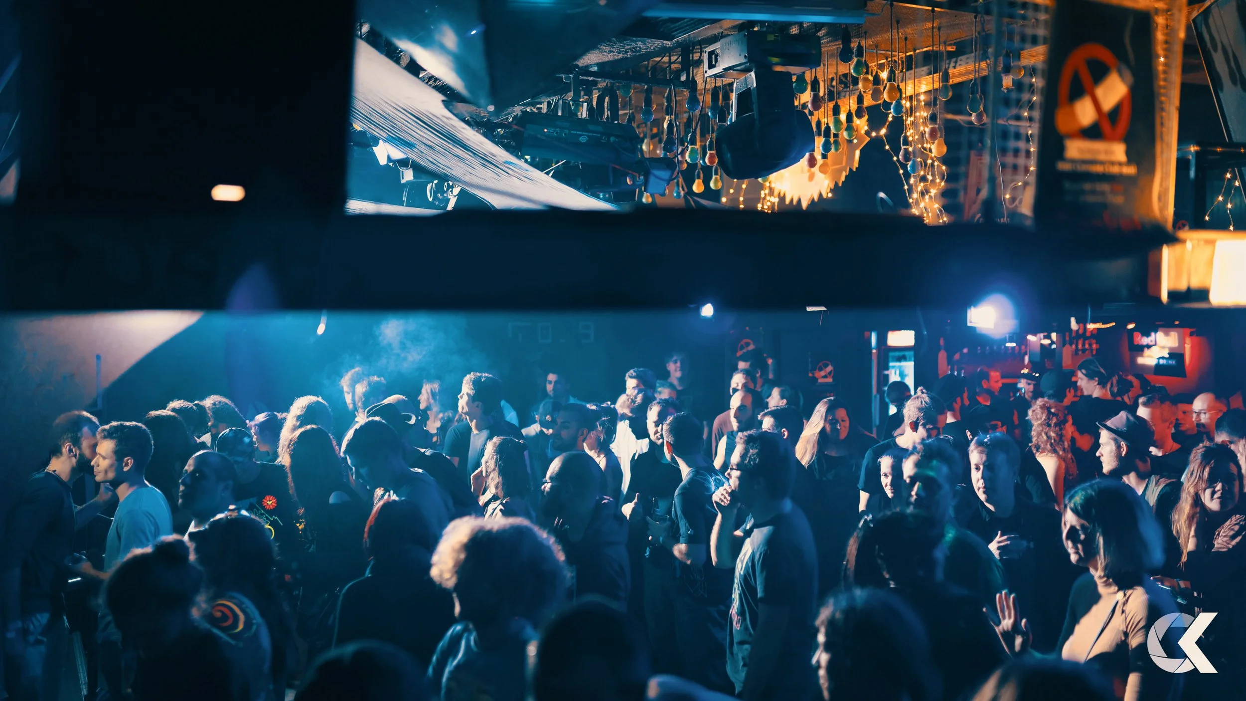 Crowd of people dancing and socializing inside a nightclub with blue and orange lighting, decorated with hanging ornaments and string lights.