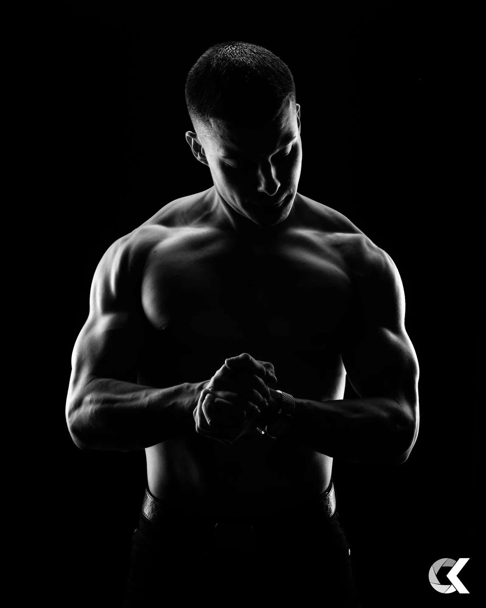 A black and white photo of a shirtless man with muscular build looking down and adjusting his watch, against a dark background.