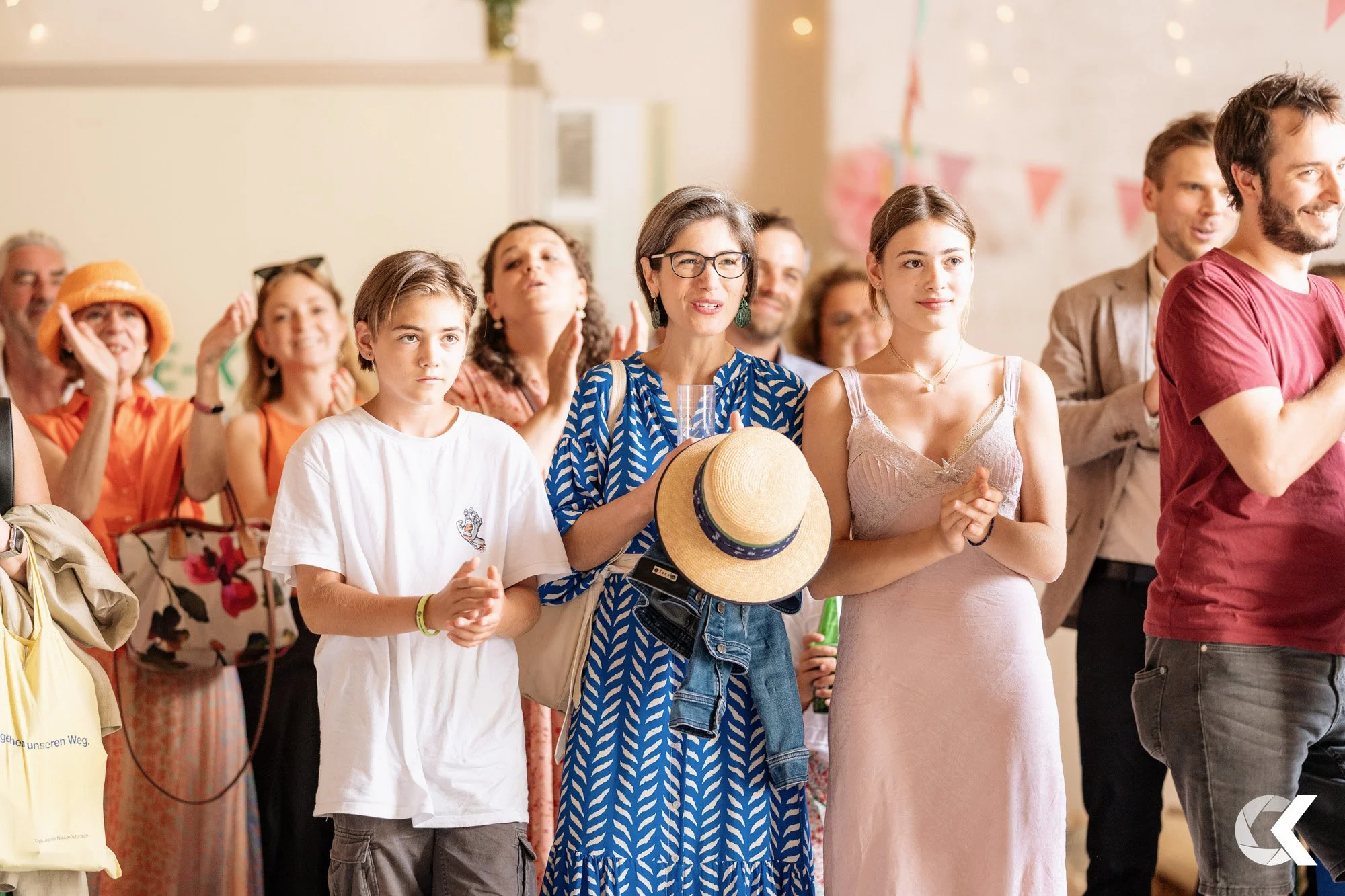 Group of people at an indoor event, some clapping and smiling, including a woman in glasses holding a straw hat, a teenage girl in a pink dress, and a boy in a white shirt.
