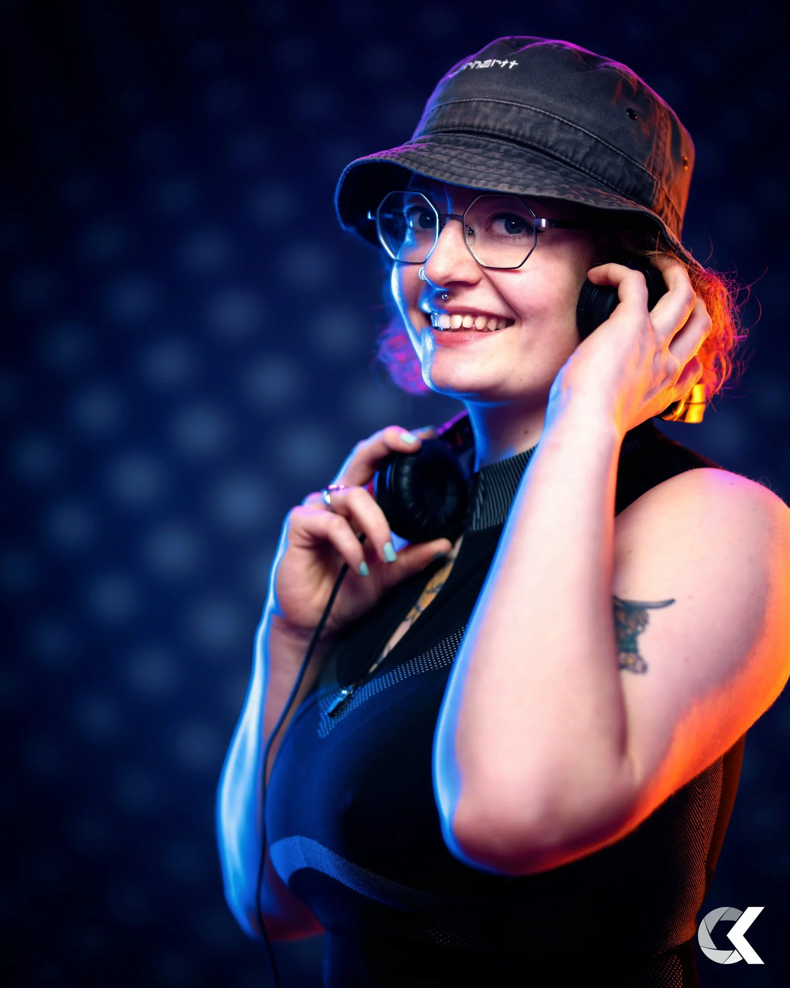 A smiling woman with glasses and tattoos, wearing a bucket hat and headphones, standing against a dark background with colorful lighting.