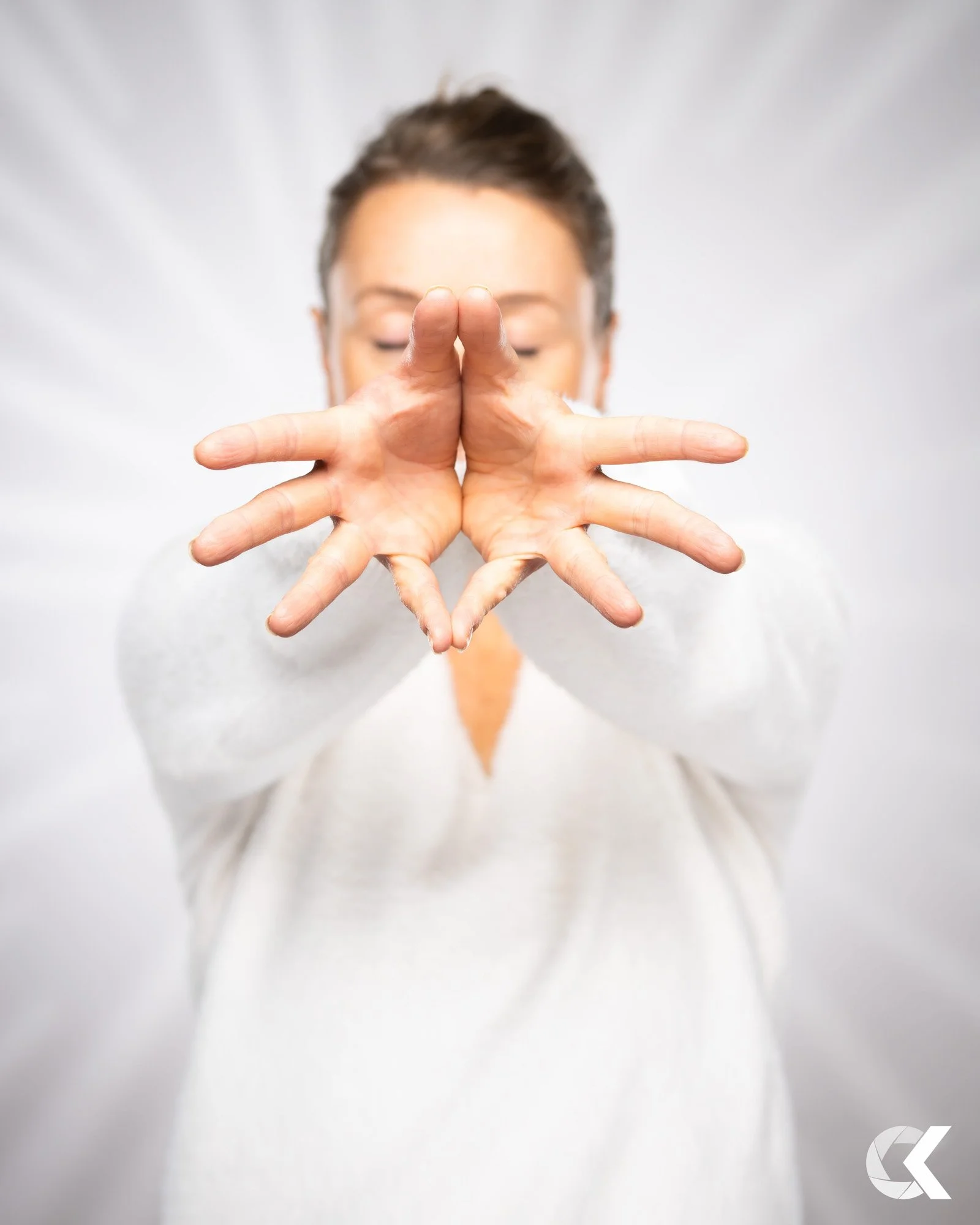A woman with short brown hair wearing a white sweater, holding her hands out in front of her with fingers spread apart, facing the camera, with a neutral background.