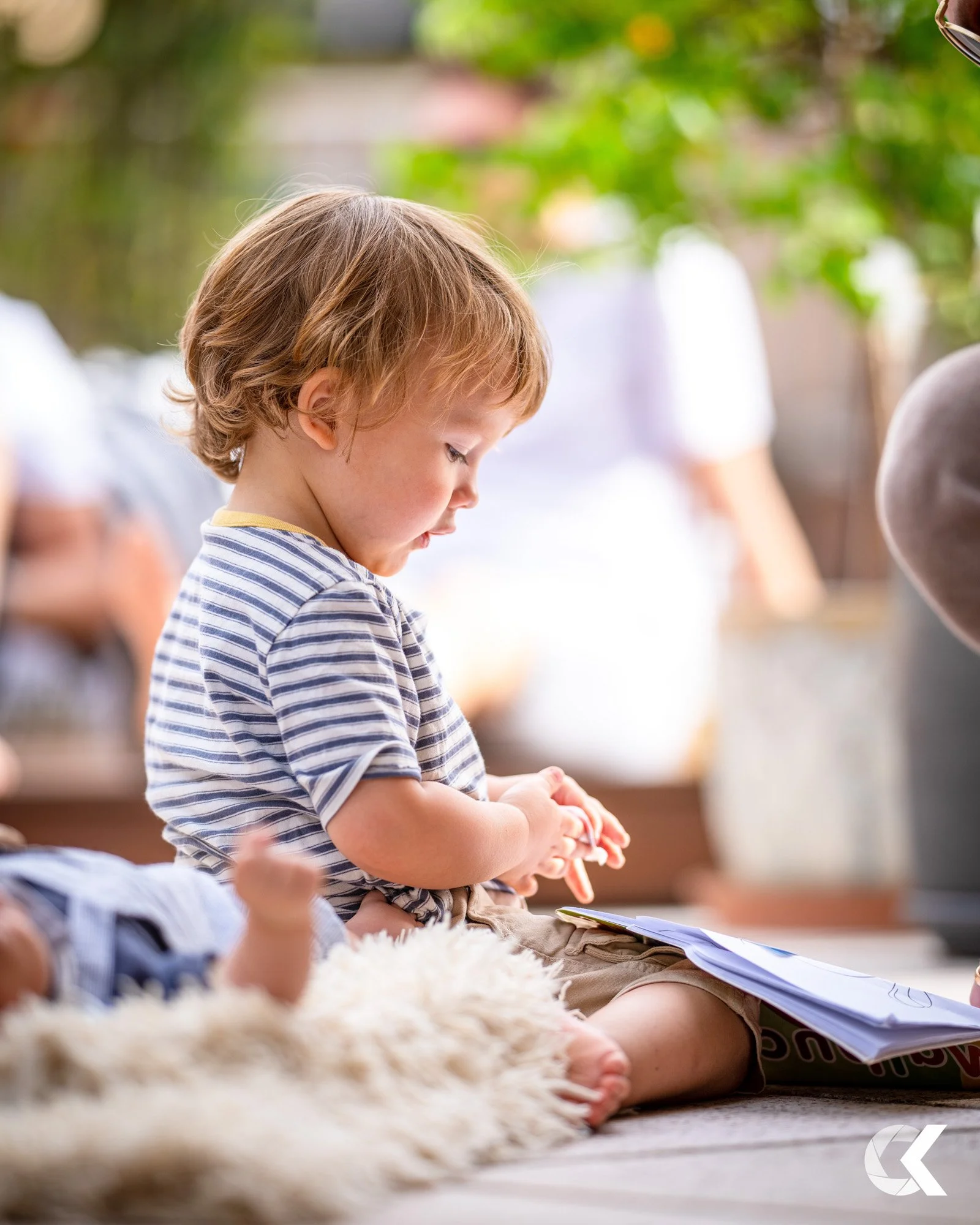 A young boy sitting on a rug outdoors, looking at a piece of paper he is holding with both hands, with a fluffy rug beside him and blurred greenery and people in the background.