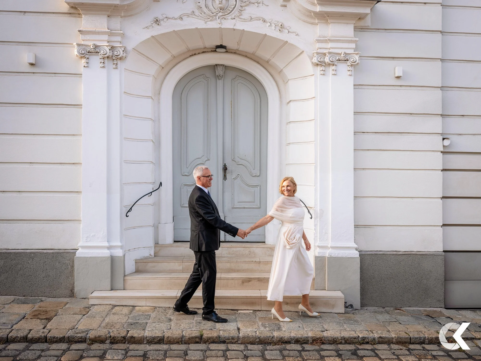 A smiling woman in a white dress and high heels holding hands with a man in a black suit outside a white building with large double doors and cobblestone ground.