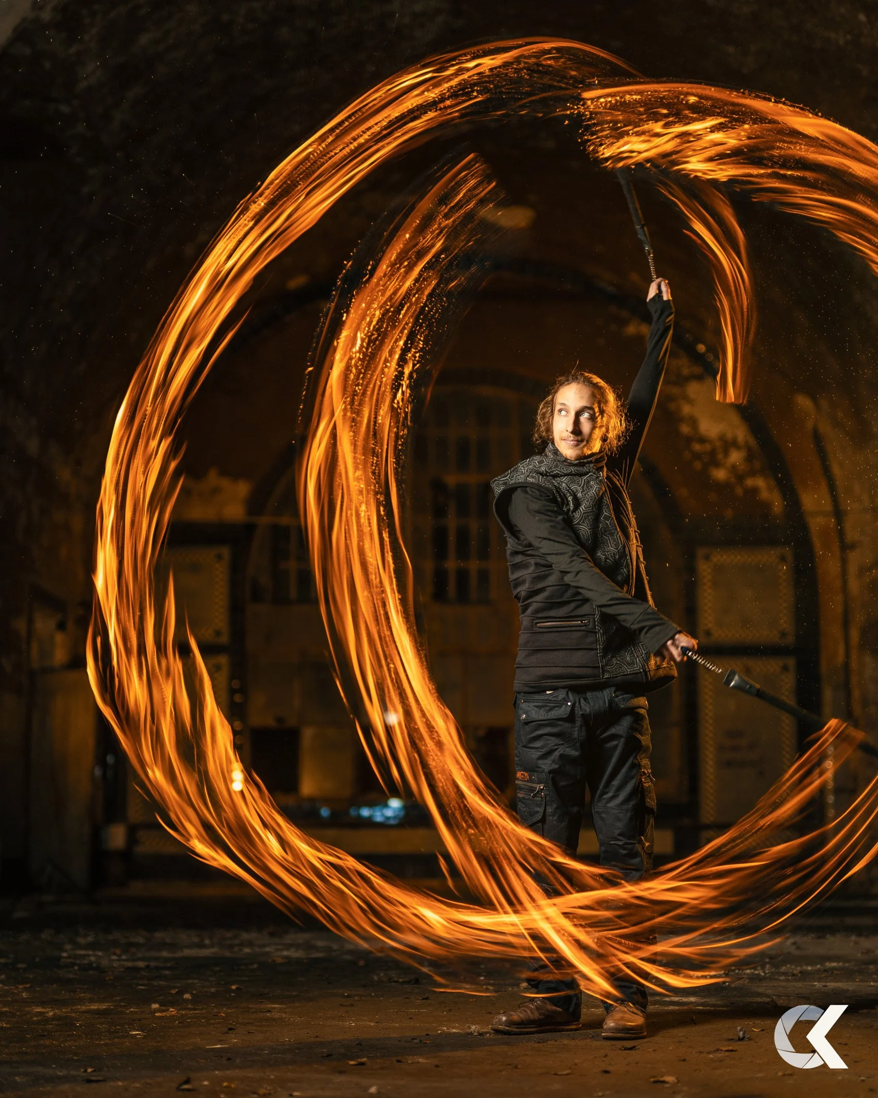 A person performing fire spinning with two fire poi, creating circular fire trails in an indoor setting at night.