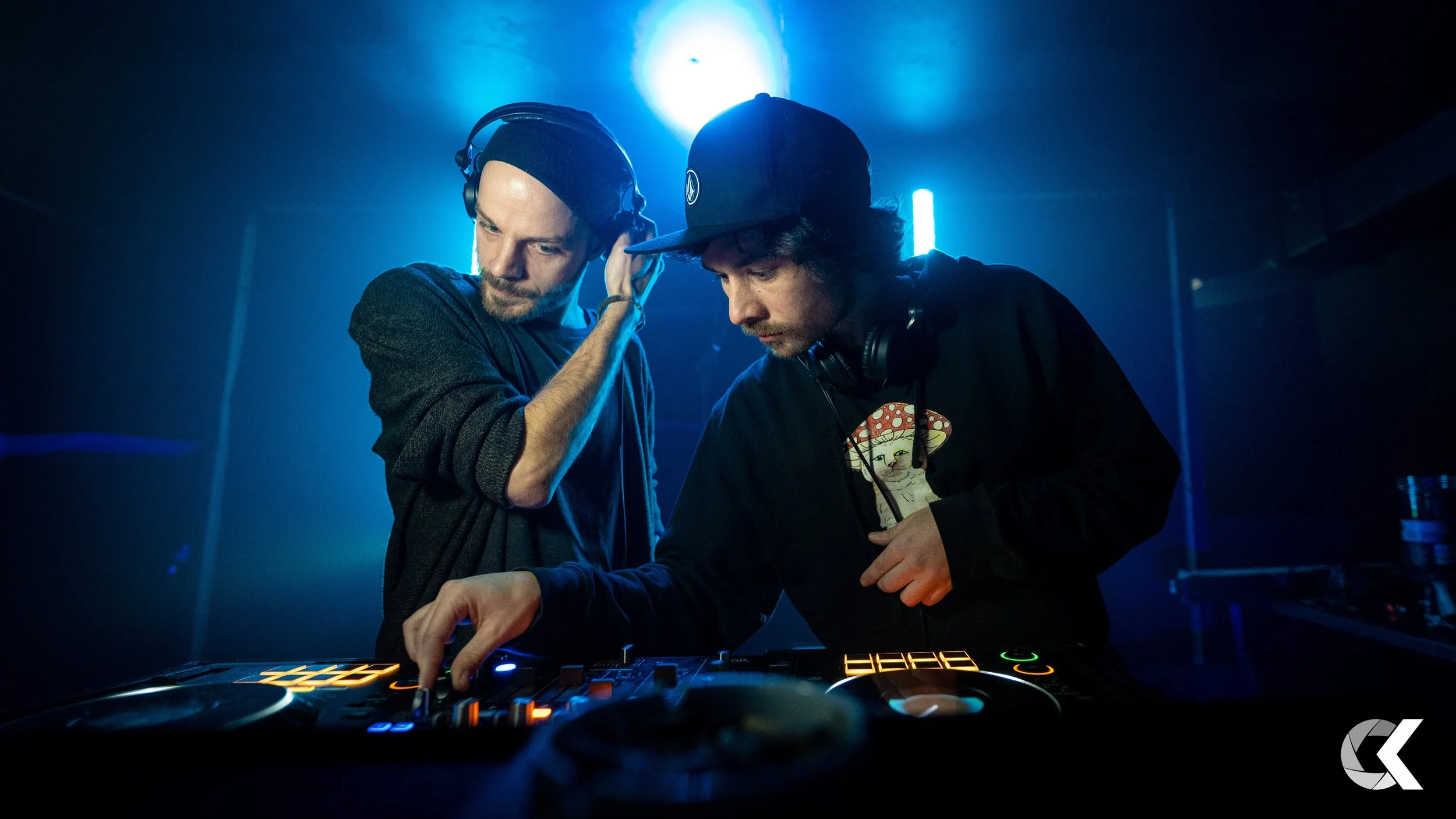 Two DJs in a dark club with blue lighting, one adjusting headphones and the other using a DJ controller.