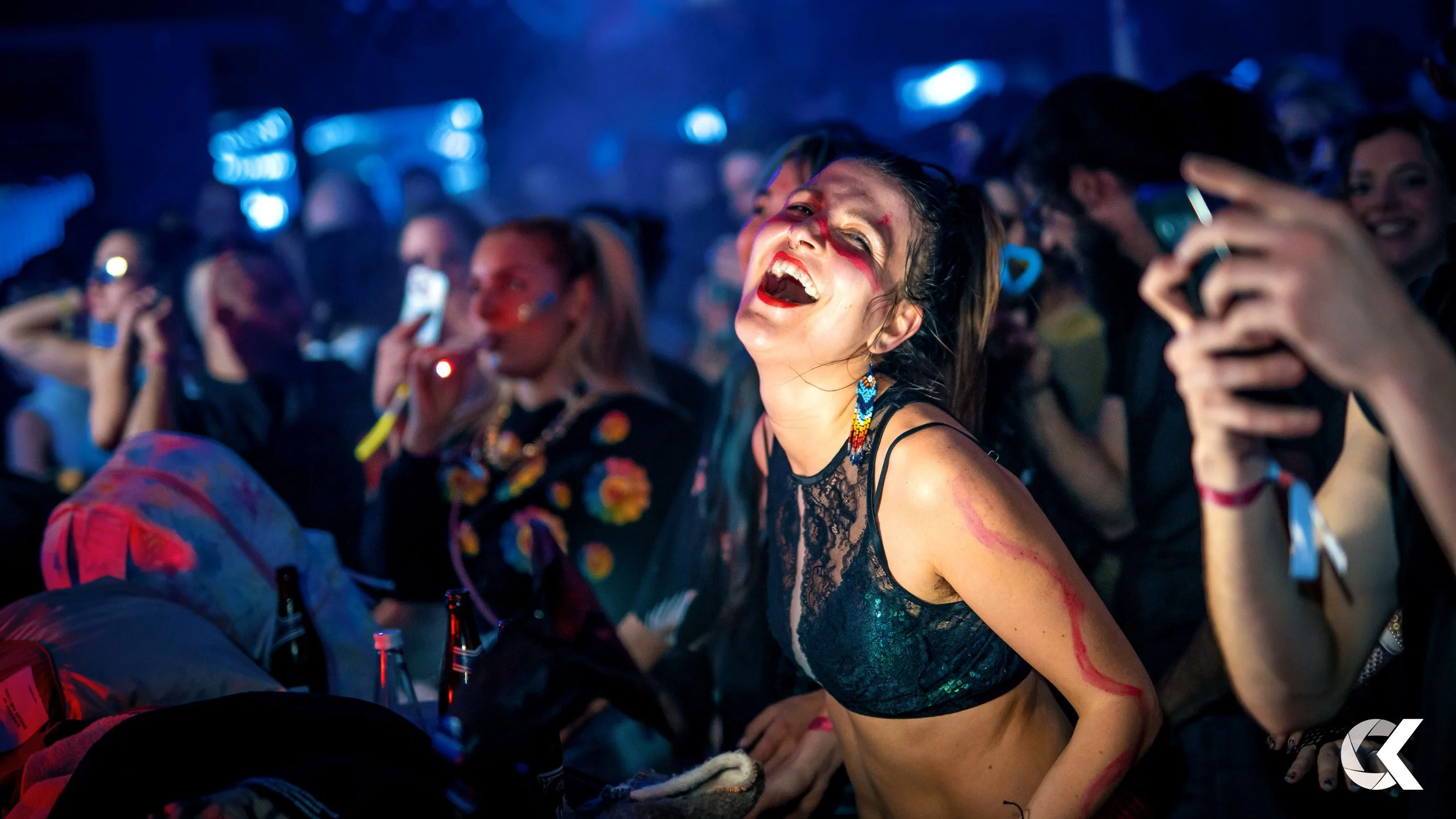 A young woman at a concert or party with face paint, earrings, and revealing top, laughing and enjoying herself surrounded by other people taking photos and videos.