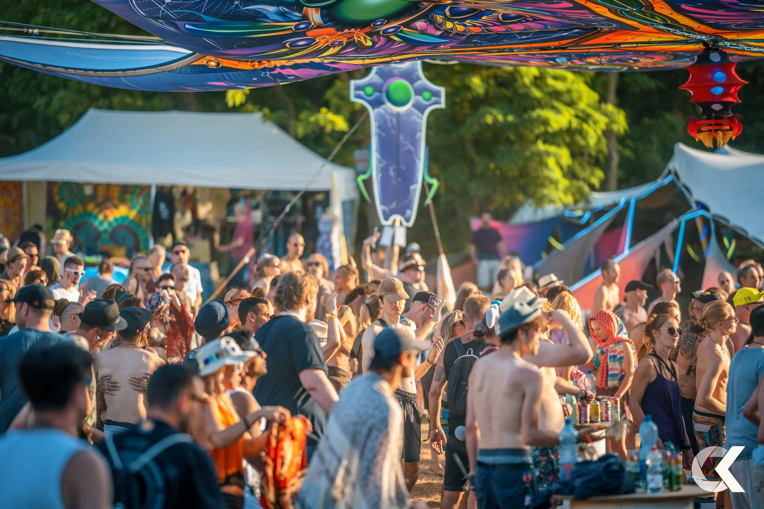 Crowd of people at an outdoor music festival during the daytime, under hanging colorful decorations and tents, with trees in the background.