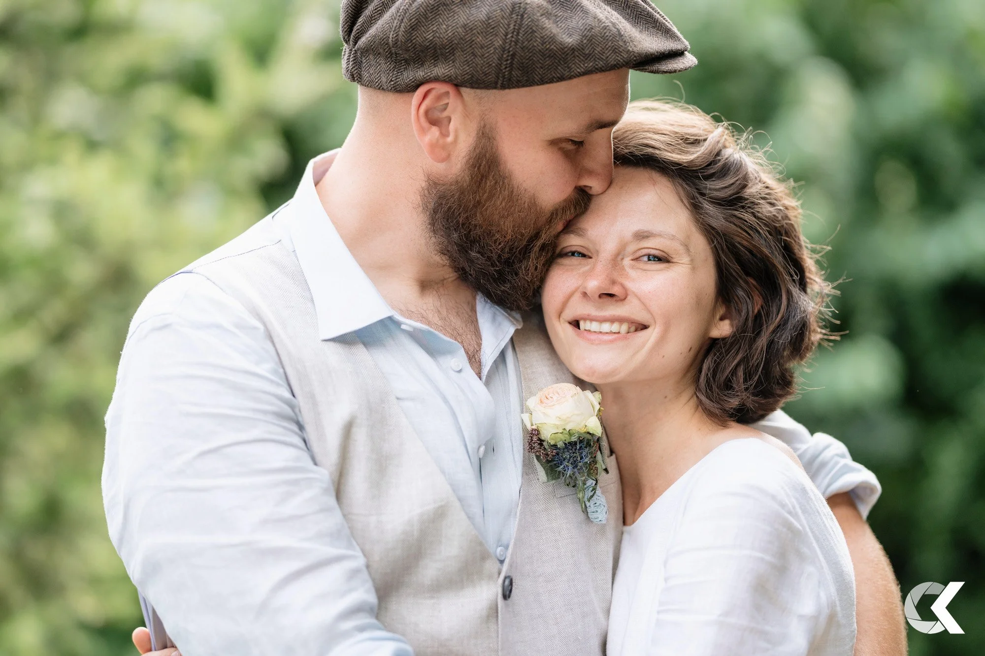 A man and woman embrace outdoors, smiling happily. The man kisses the woman's forehead as she wears a white dress and has short, wavy hair. The man has a beard, mustache, and wears a brown cap and a light gray vest over a white shirt. The woman has n