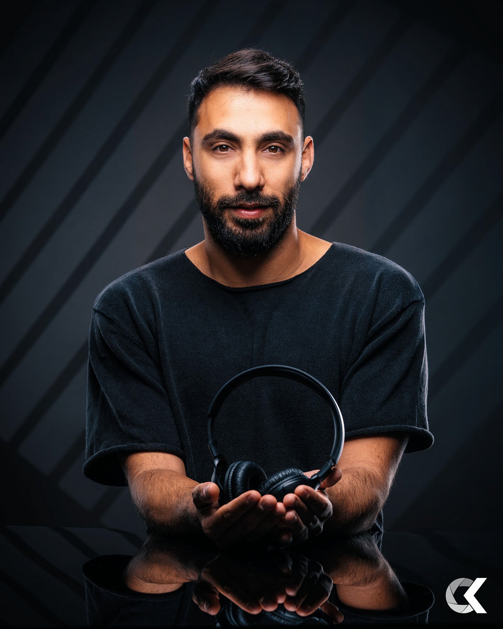 A man with a beard and short dark hair holds a pair of black headphones in front of a dark, geometric background.