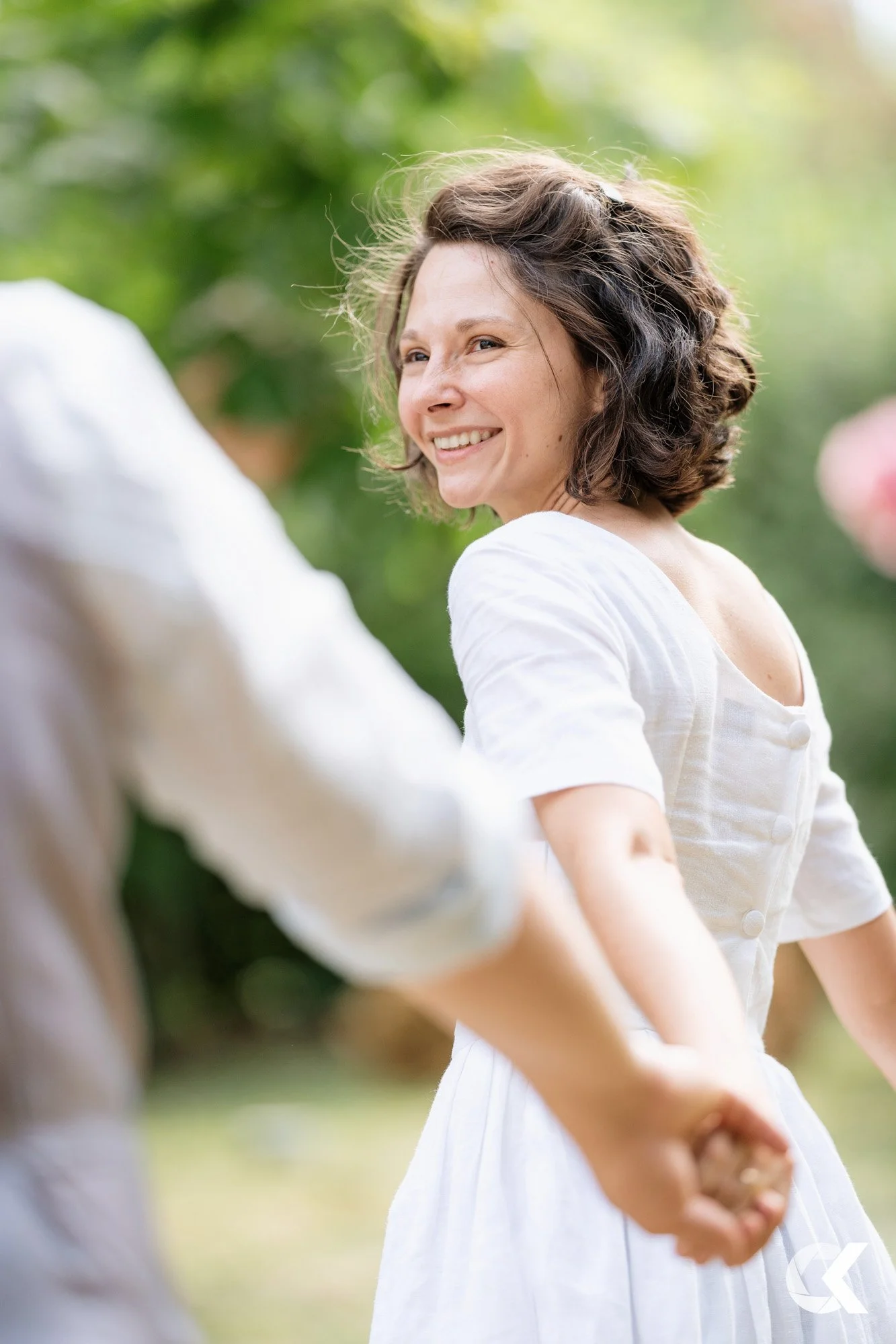 Woman with curly brown hair smiling and holding hands with a person, outdoors with green foliage in the background.