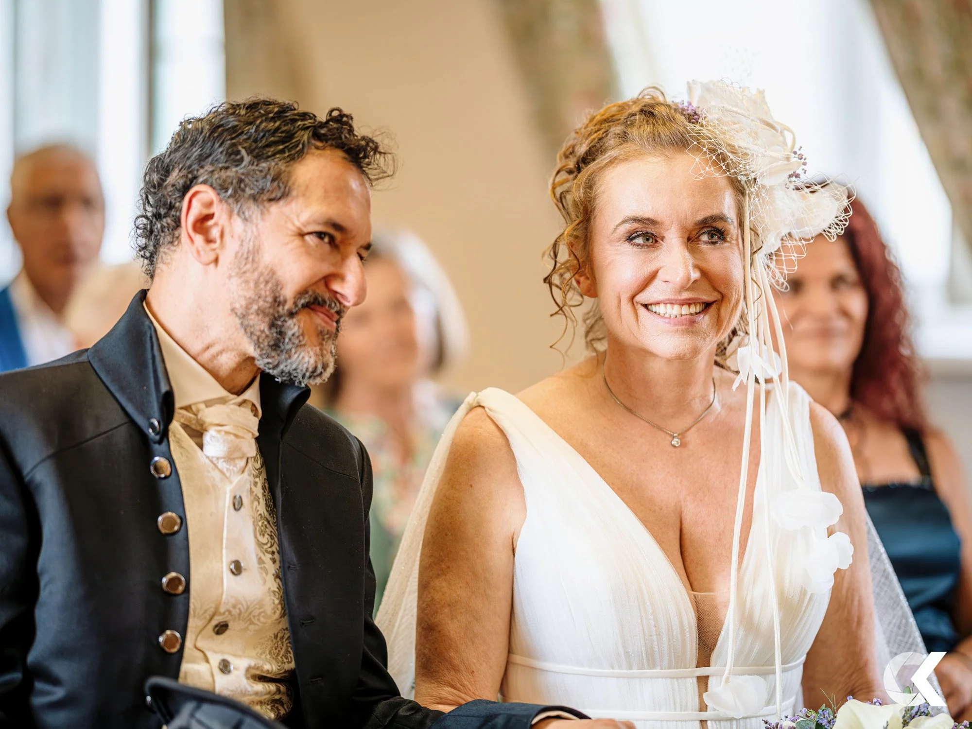 A woman in a wedding dress with a large white flower and netting on her head, smiling, sitting during a wedding ceremony. Next to her is a man in a vintage-style black suit and an ivory vest, also sitting, with other guests in the background.