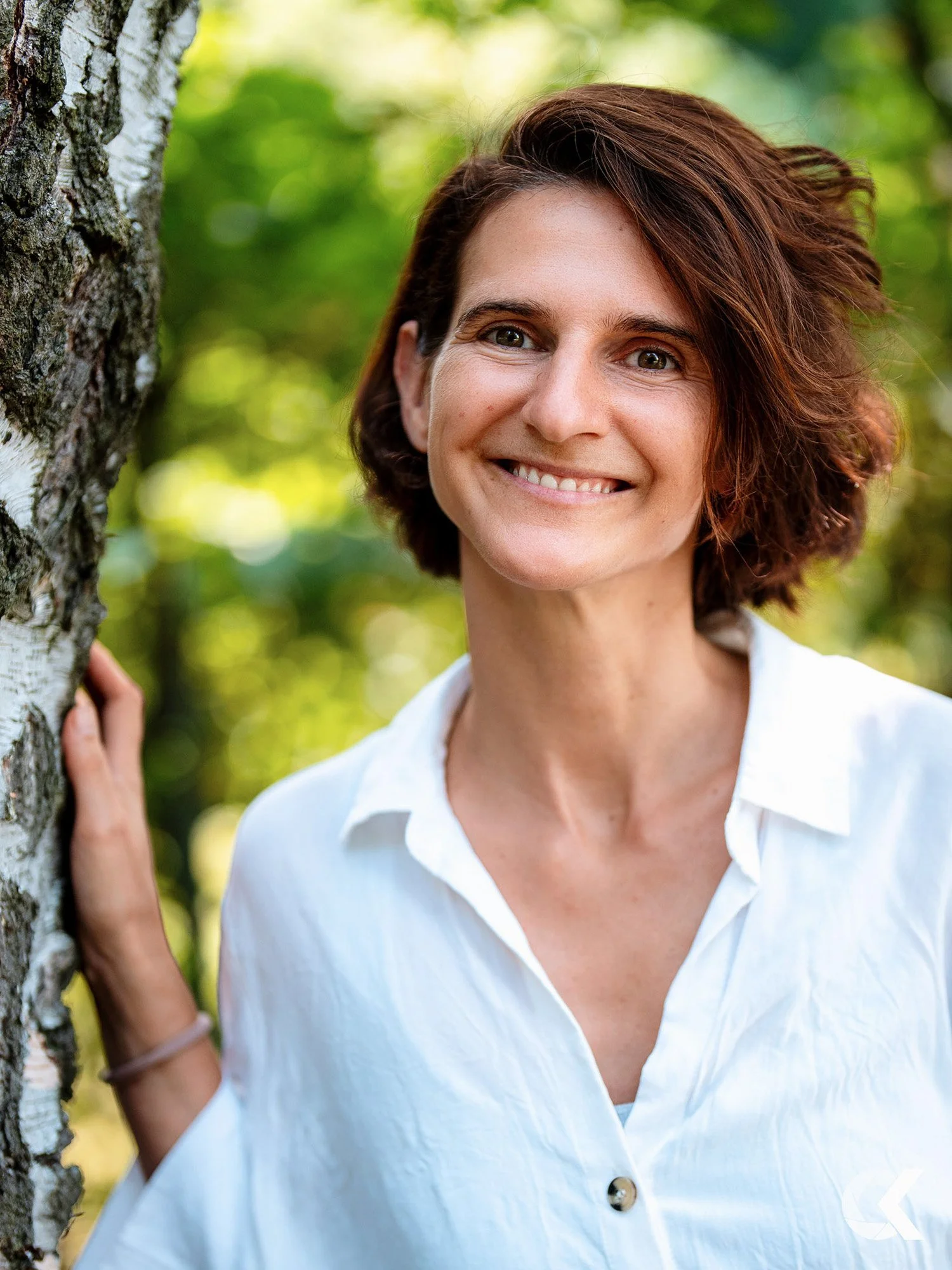 A woman with short brown hair and a white shirt smiling outdoors in a green, leafy environment.
