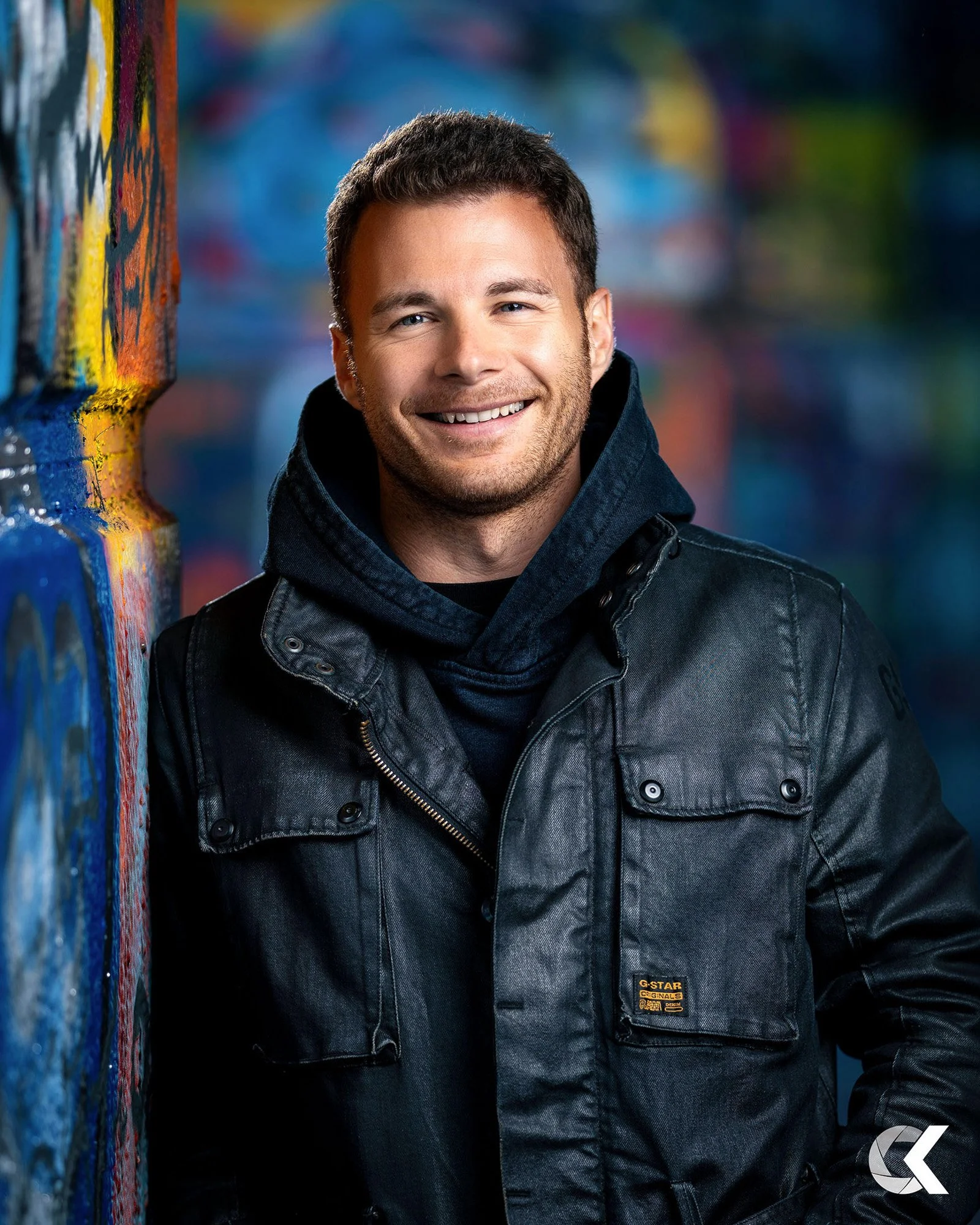 A smiling man with short dark hair, wearing a black leather jacket and hoodie, standing beside a colorful graffiti wall.