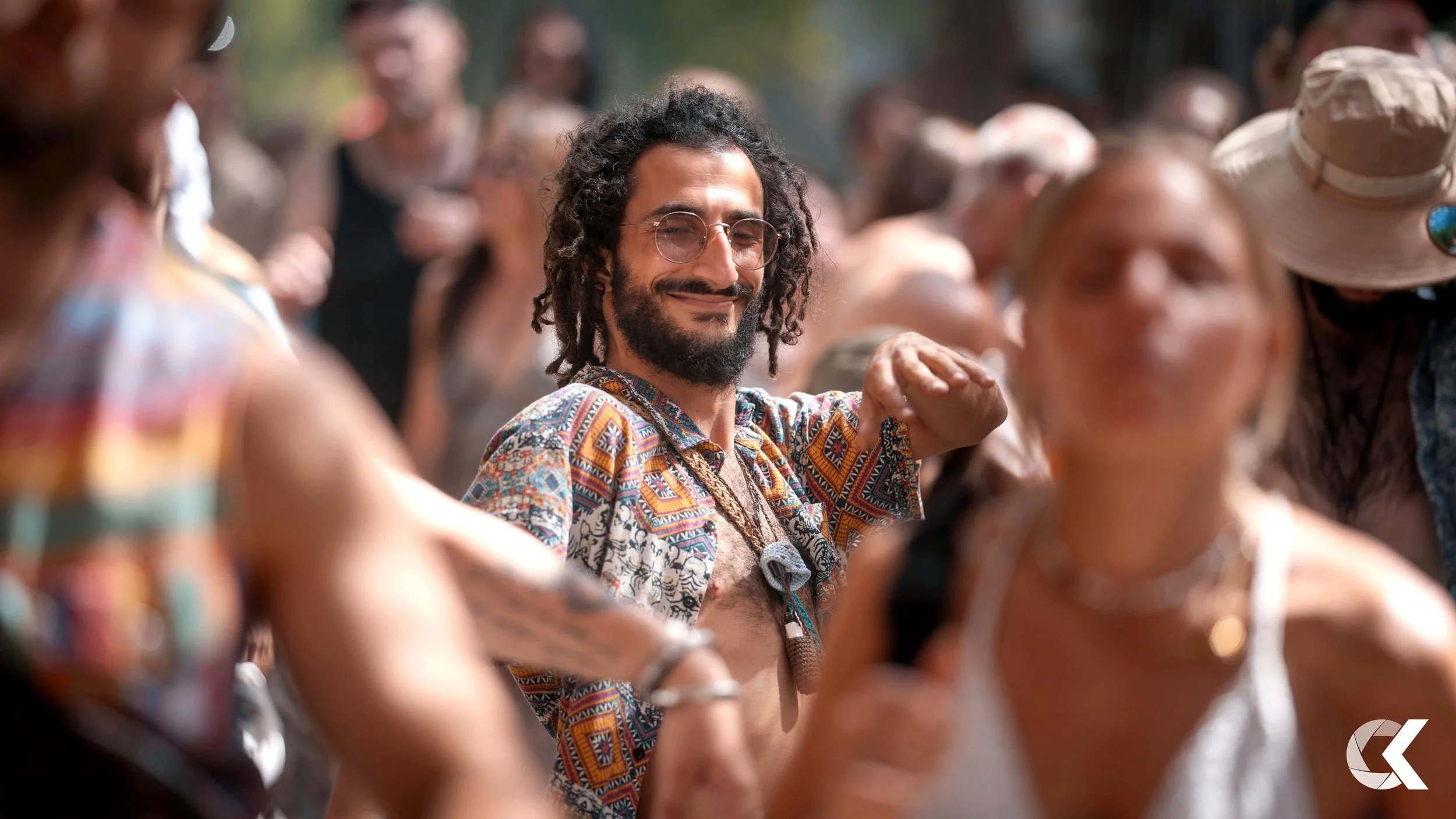 A group of people dancing outdoors at a festival, with a man in the center smiling and wearing a colorful, patterned shirt.