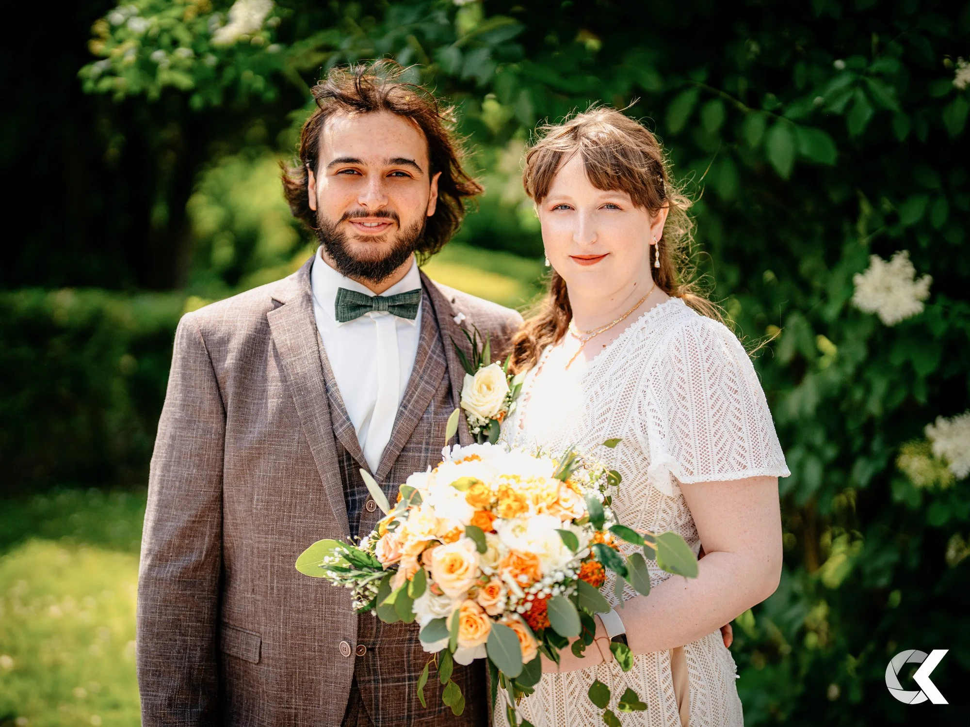 A bride and groom standing outdoors in a garden, smiling at the camera, with the bride holding a large bouquet of flowers.