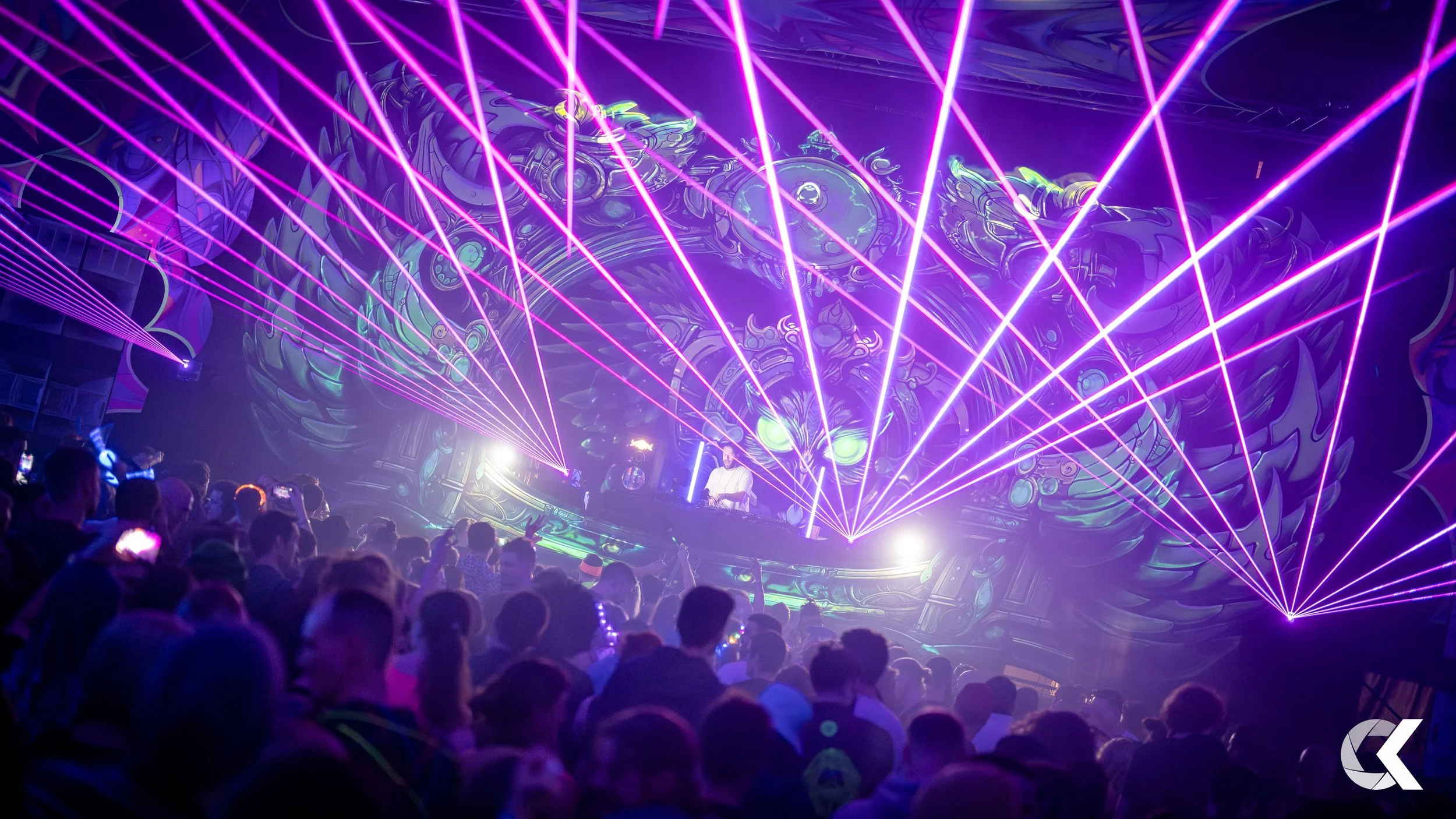 A DJ performs at a nightclub with laser lights and vibrant visuals on the wall behind him, and a crowd of people enjoying the music.