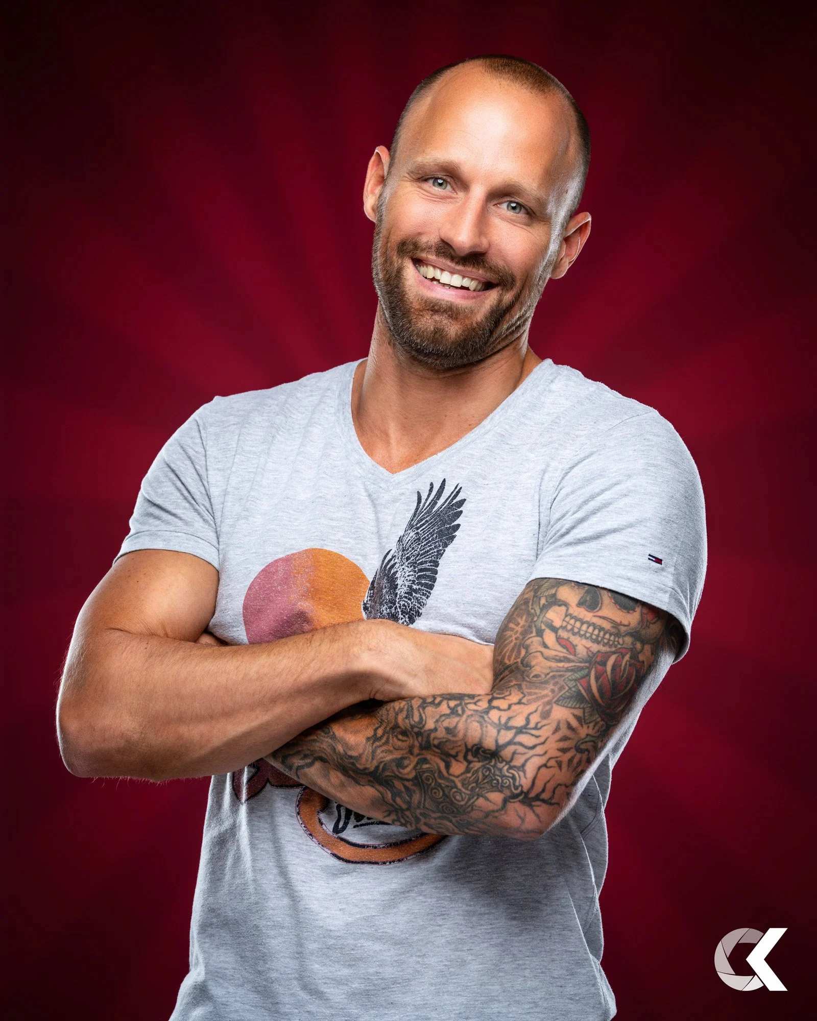A smiling man with a beard and tattoos on his right arm, wearing a light gray T-shirt, stands with arms crossed against a red background.