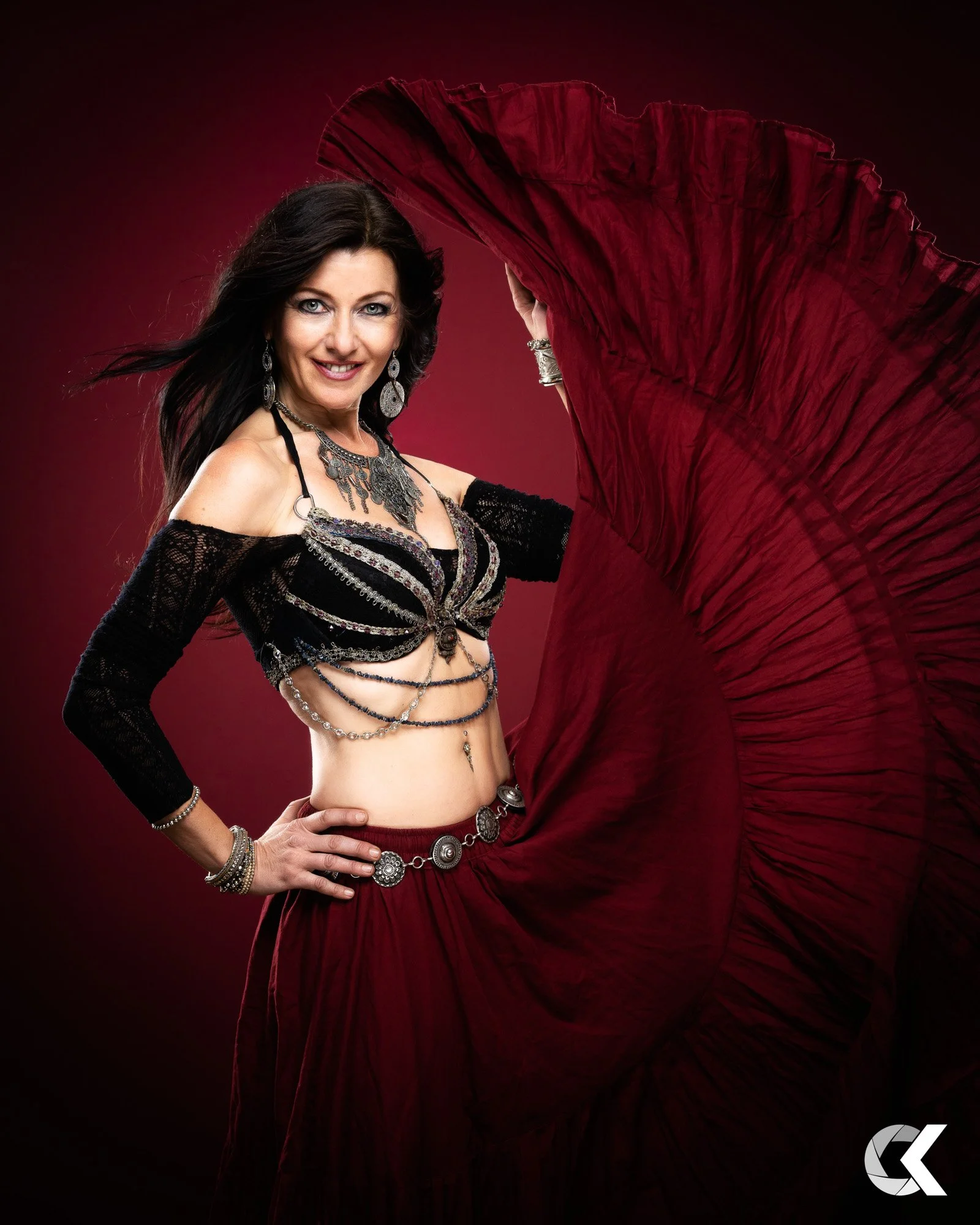 A woman with long dark hair, smiling, wearing elaborate jewelry and a black top, holding a large red fabric in front of a red background.