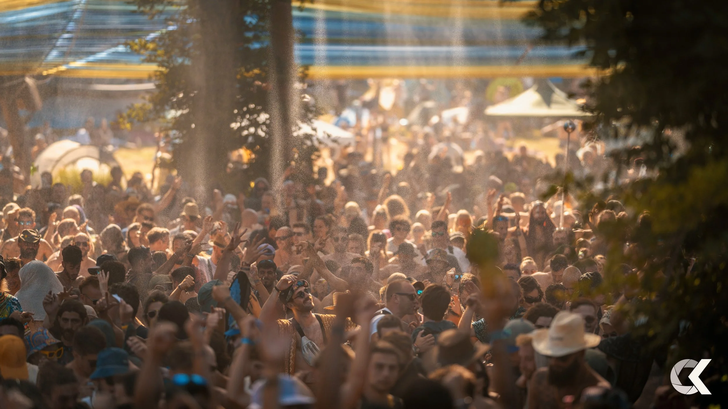 Crowd of people at an outdoor festival with trees and tents in the background, some people are taking photos and raising their hands in the air.