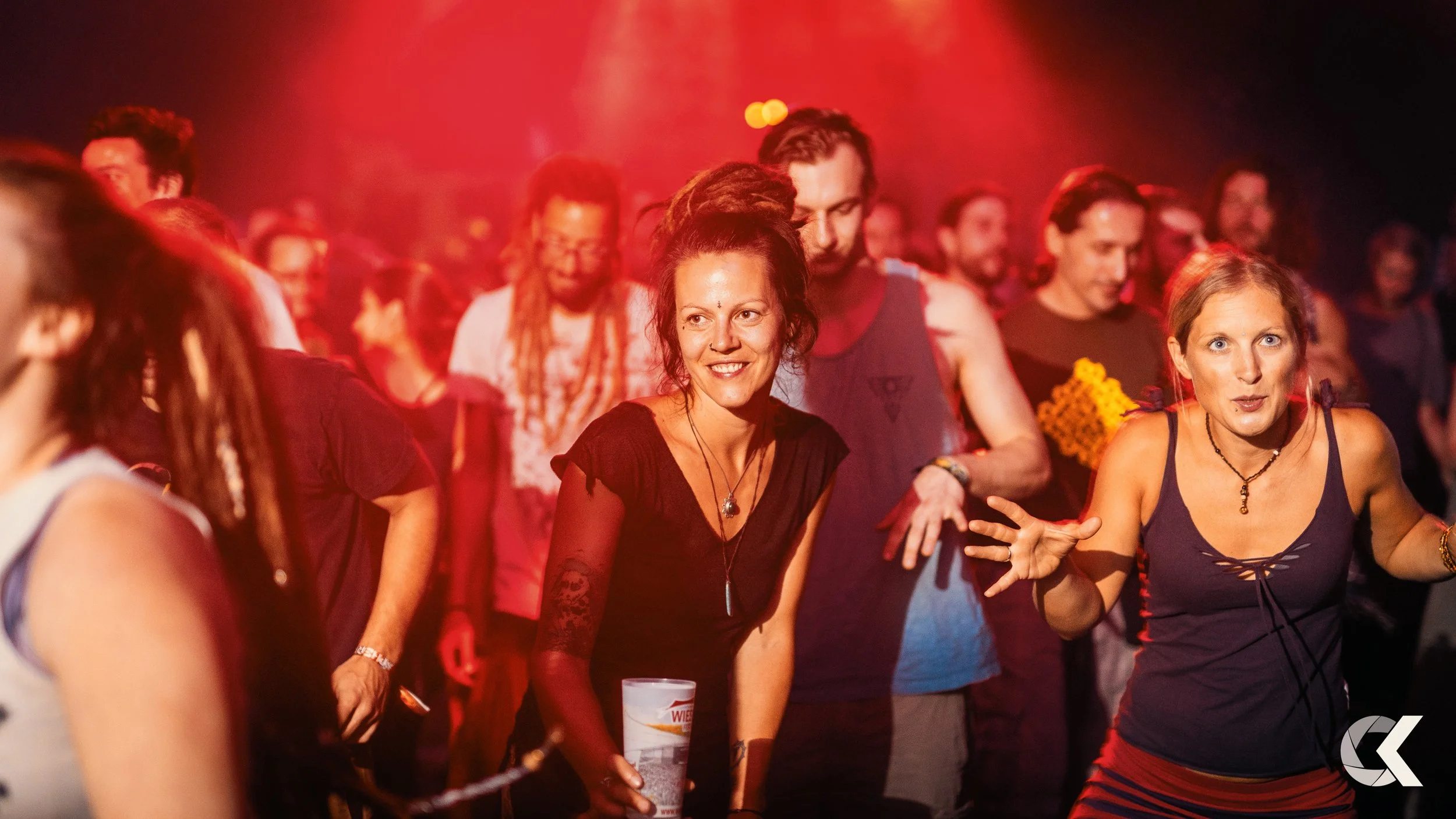 People dancing at a party with red lighting, two women in the foreground dancing and holding drinks, surrounded by other partygoers.