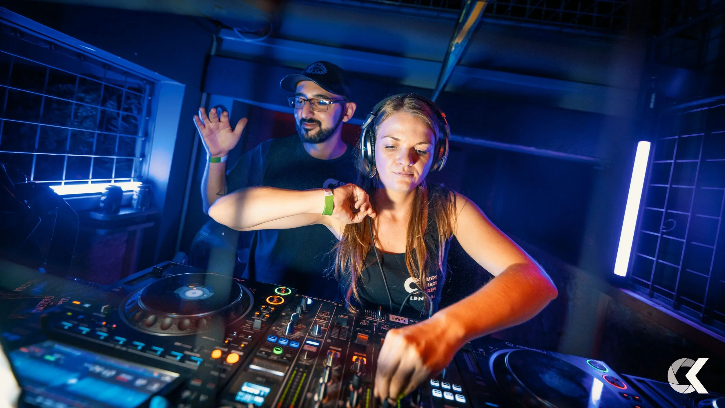 Female DJ wearing headphones and adjusting controls on a DJ mixer in a nightclub setting, with a male DJ behind her.