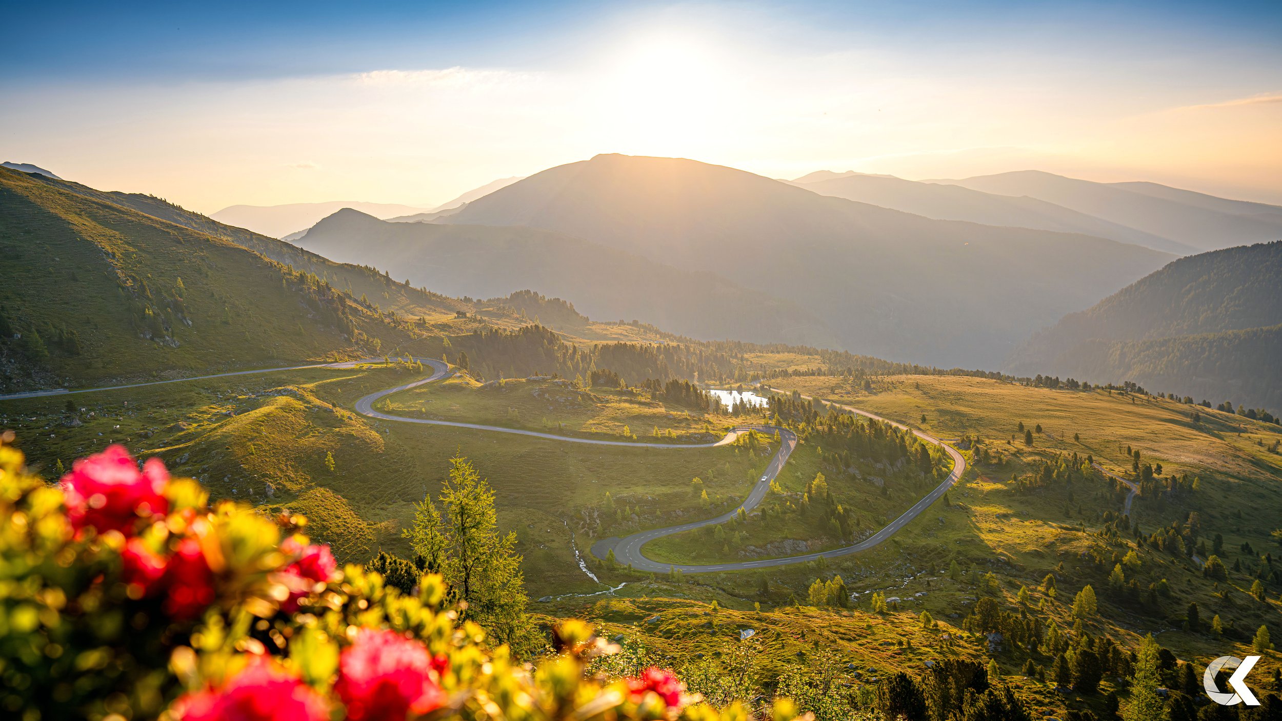Sunset over a winding mountain road with green hills, trees, and a distant lake.