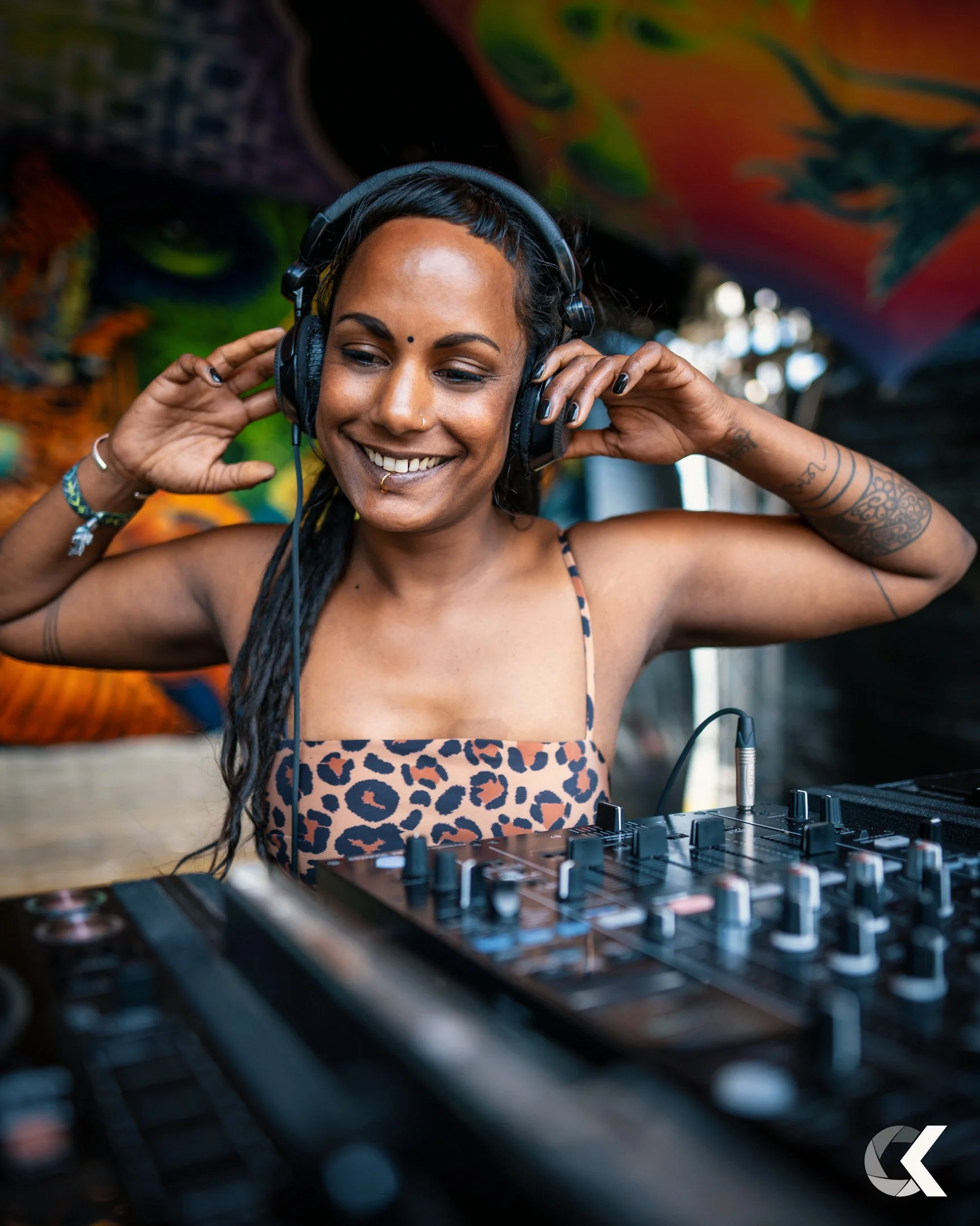 A smiling woman with long dark hair, tattoos, and a nose piercing is wearing headphones and standing in front of a DJ mixer, adjusting her headphones with her hands. The background is colorful and artistic, suggesting a lively and creative environmen