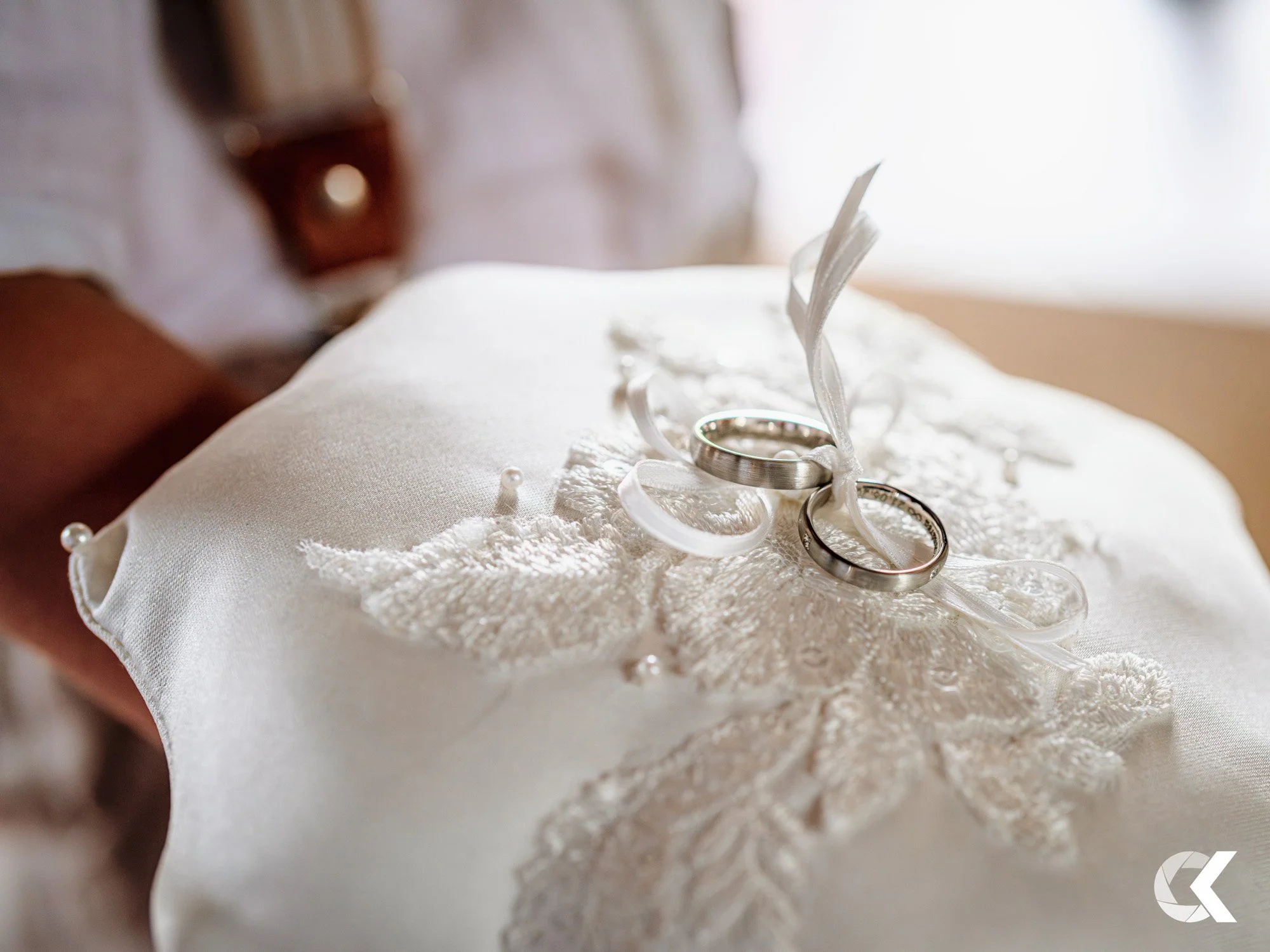 Two wedding rings on lace pillow with satin ribbons and pearls, held in a person's hand.