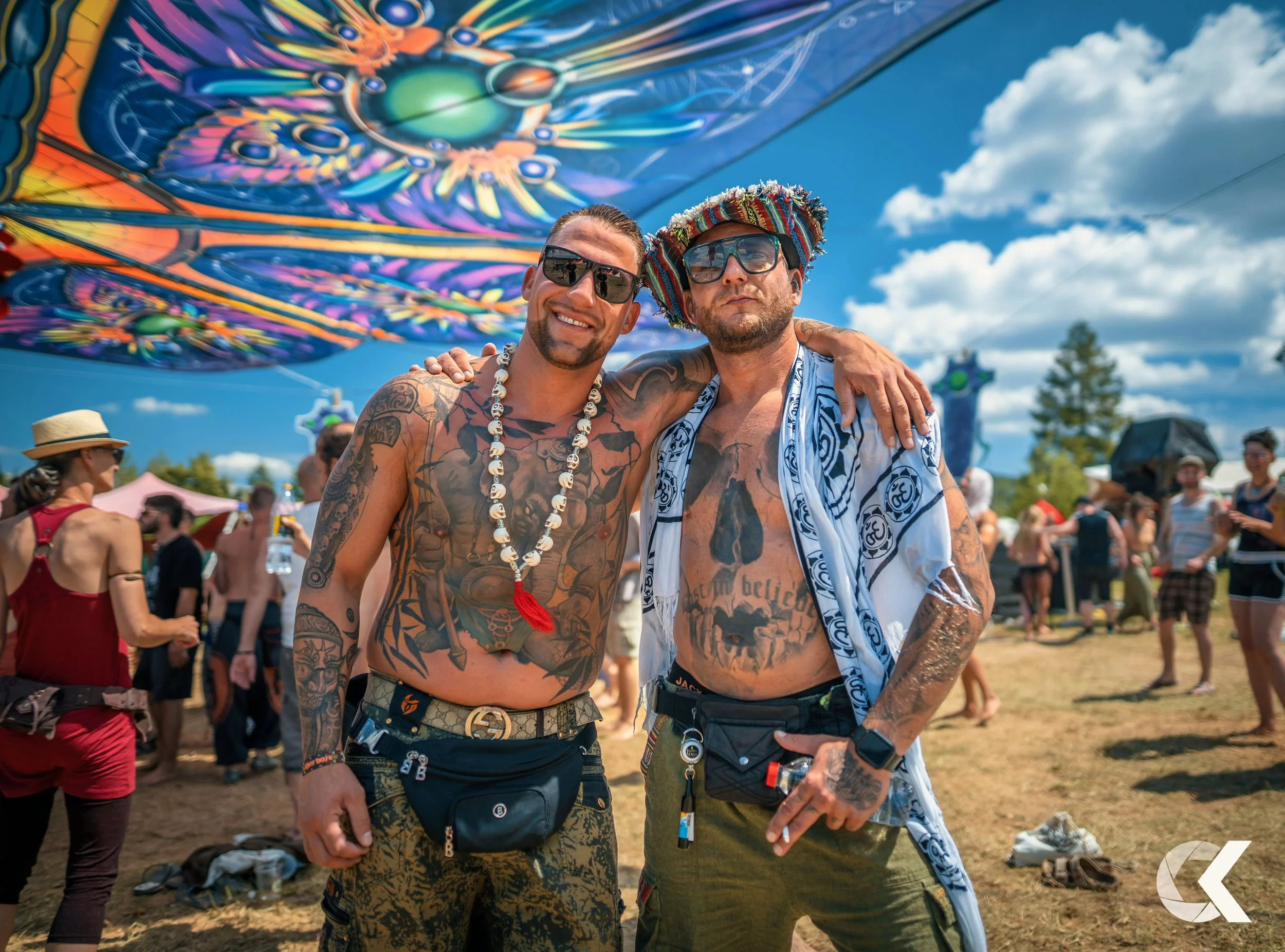 Two tattooed men with sunglasses and accessories, standing outdoors at a festival, with colorful umbrellas overhead and crowds in the background.