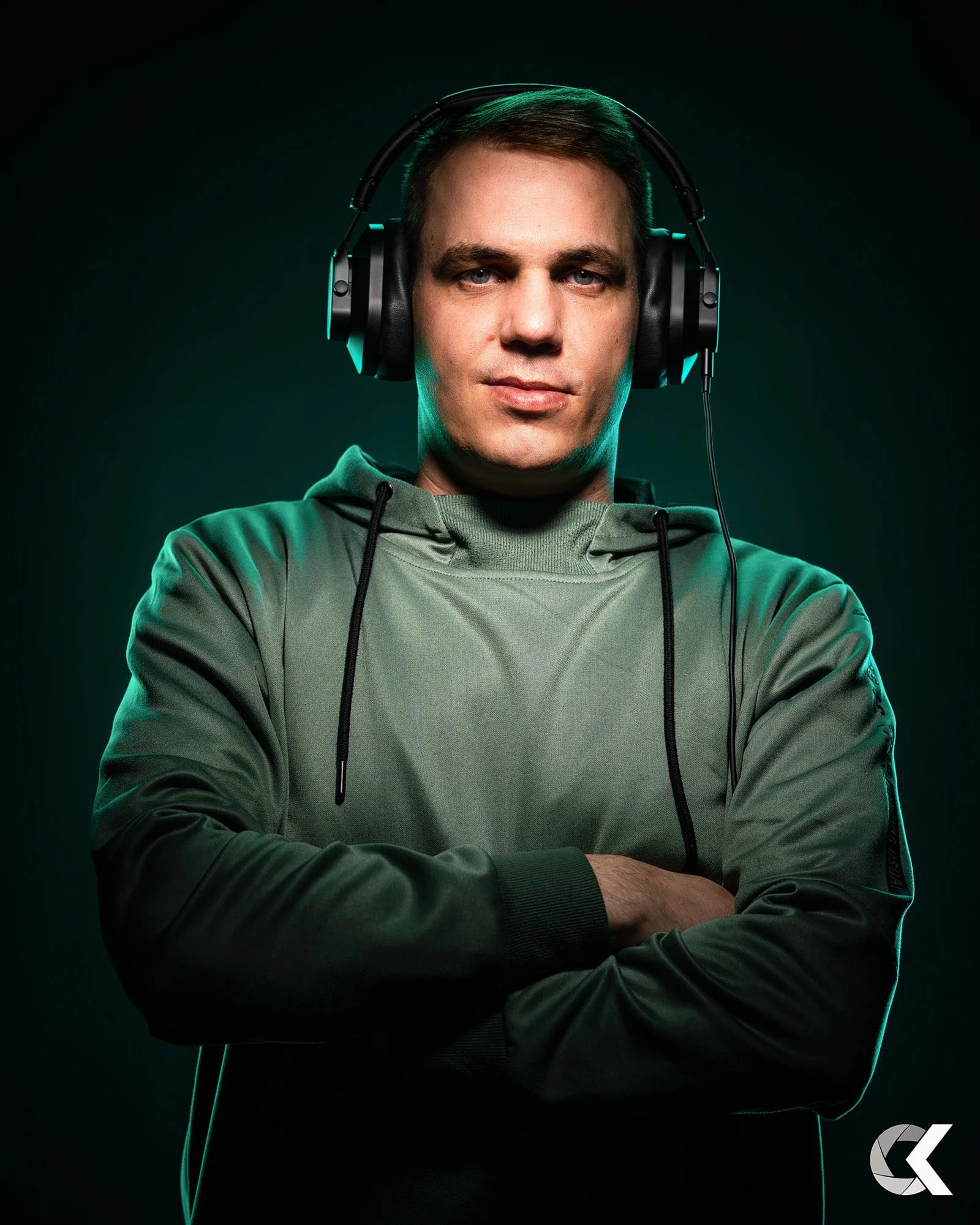 Man wearing large over-ear headphones and green hoodie with arms crossed, posing in a dark studio with green lighting.
