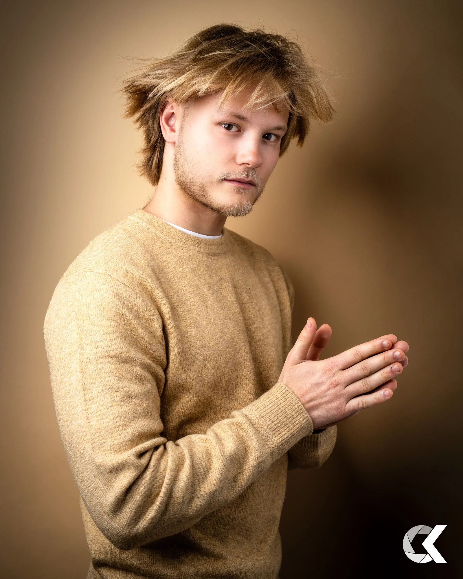 A young man with medium-length blond hair and light facial hair, wearing a beige sweater, standing against a beige background, with his palms pressed together in a prayer or greeting gesture.