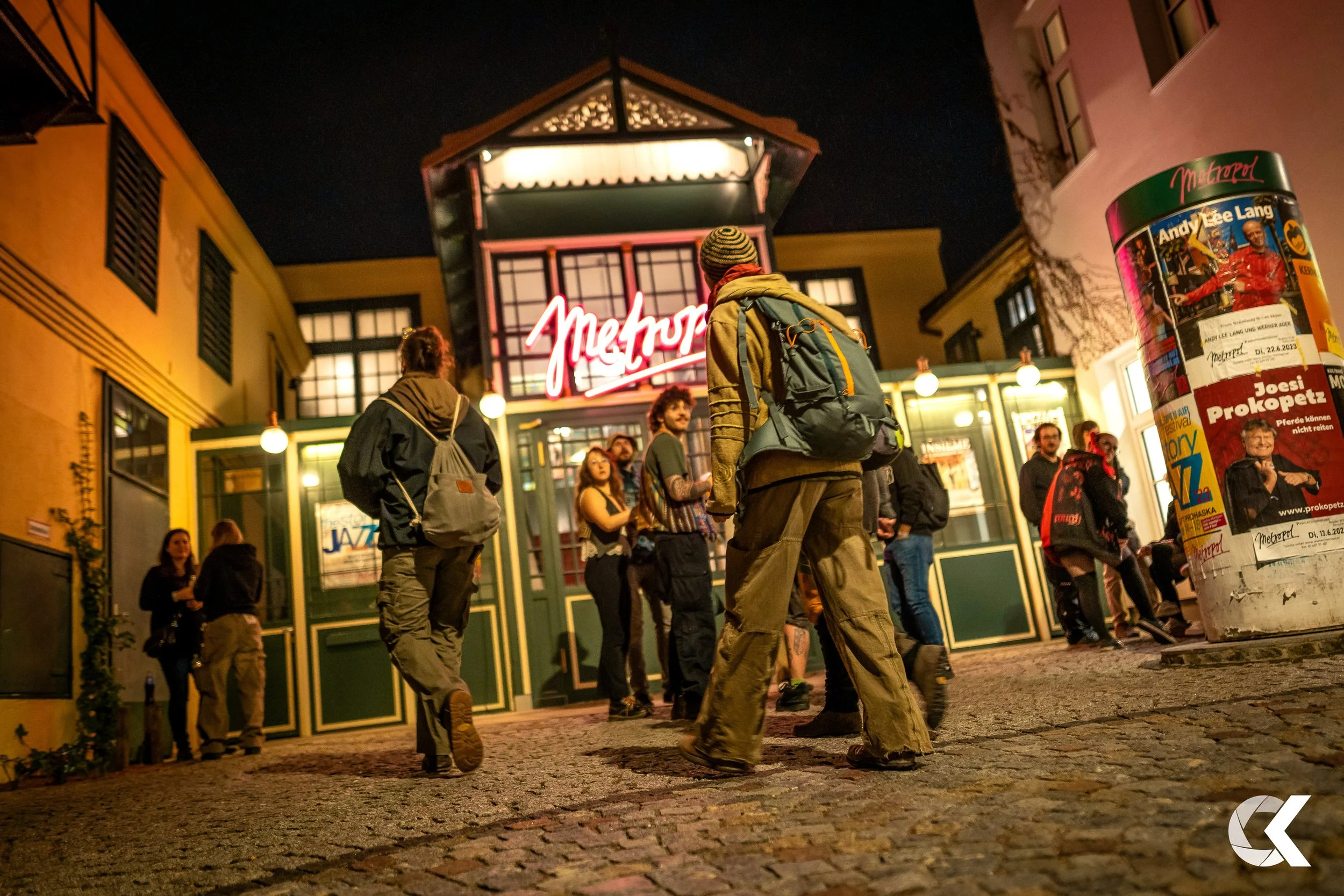People gathered outside a building with a neon sign that reads 'Metropol' at night. Some are chatting, and others are walking on cobblestone street. A large cylindrical poster with German text is visible on the right.