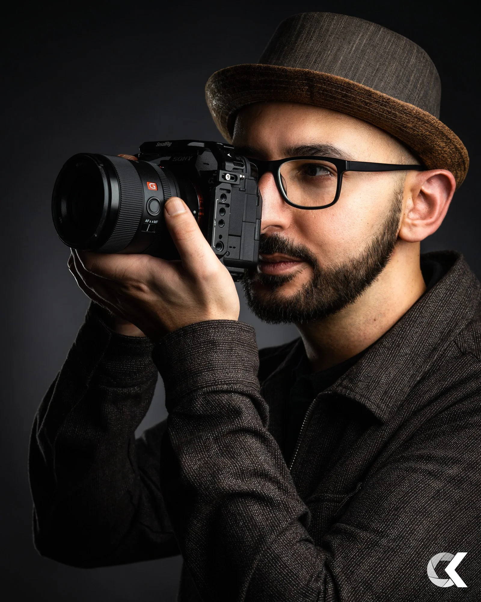 Ein Mann mit Brille und Hut, der eine Kamera hält und fotografiert, vor einem dunklen Hintergrund.